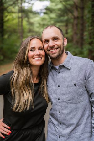 A smiling couple poses together on a forest path, woman in black dress with long hair, man in gray button-up shirt.