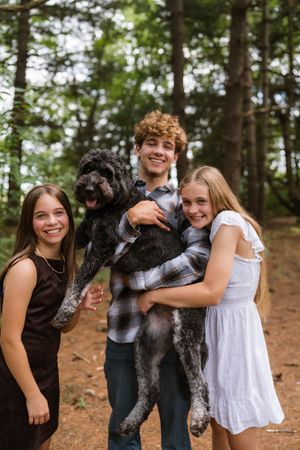 Three smiling teens pose with a black curly-haired dog in a lush forest setting.