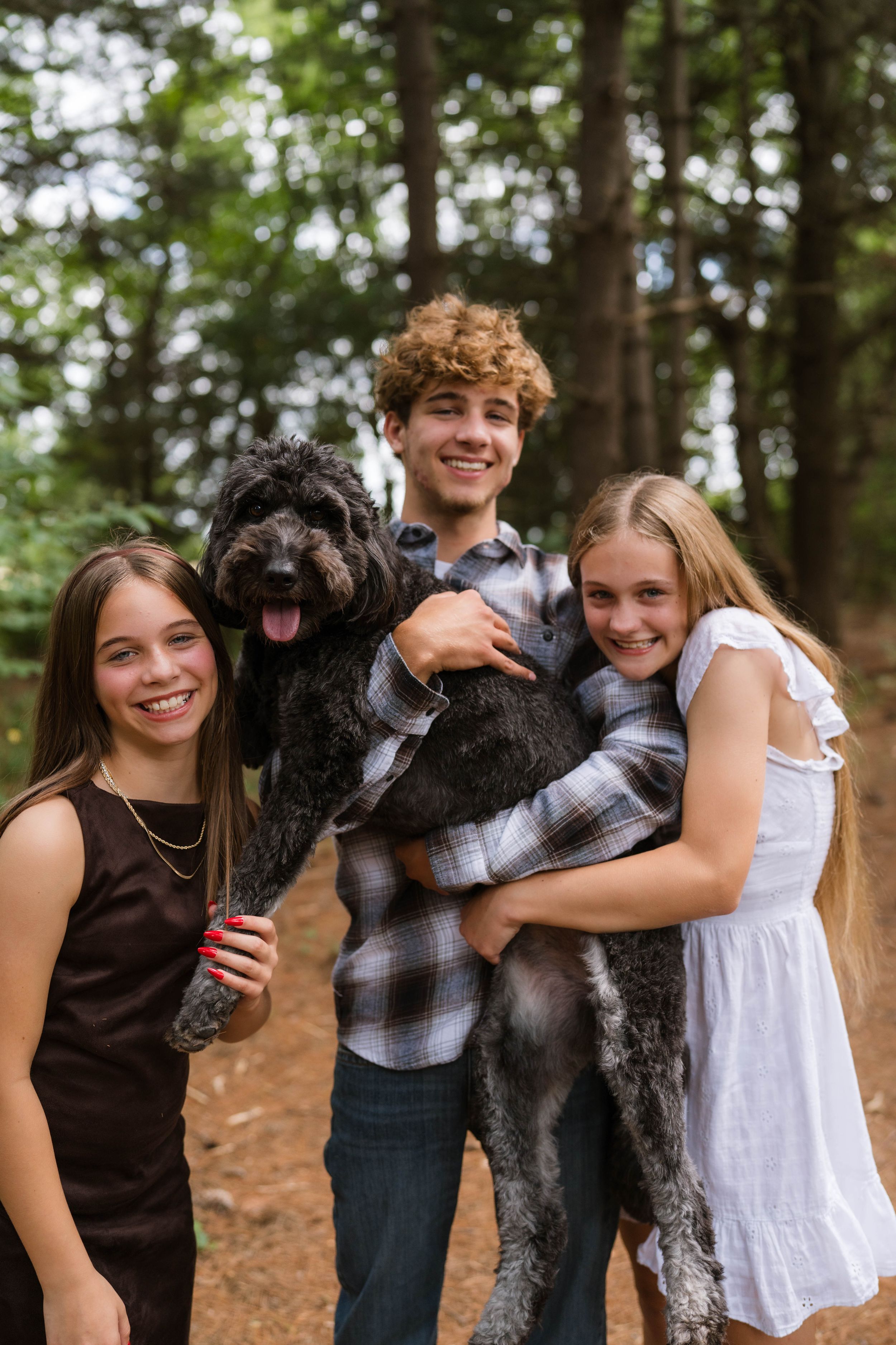 Three smiling teens hold a fluffy black dog in a sunlit forest setting.