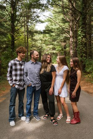 Family of five smiling together on a wooded path, dressed in casual attire, surrounded by tall pine trees.