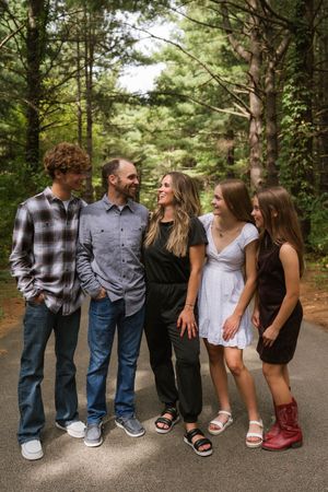 Family of five smiling and laughing together on a forest path surrounded by tall green trees.
