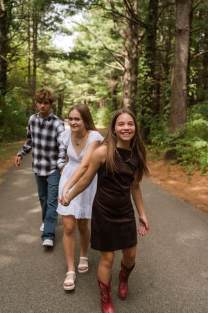 Three teenagers walking down a tree-lined path, two girls in dresses and a boy in plaid shirt and jeans.