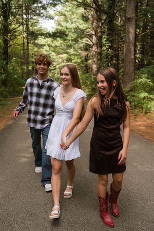 Three teens walking on a forest path, two girls holding hands in white and black dresses, boy in plaid shirt behind them.