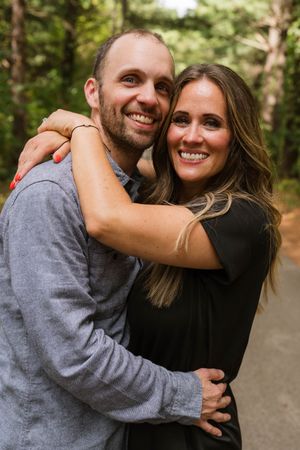A smiling couple embraces outdoors on a forest path, woman in black dress hugs man in gray shirt.
