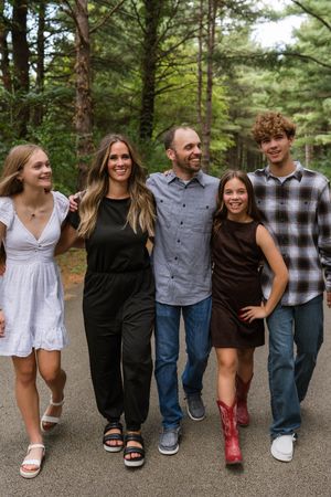 Family of five smiling and walking together on a forest path, dressed in casual outfits for an outdoor photo session.