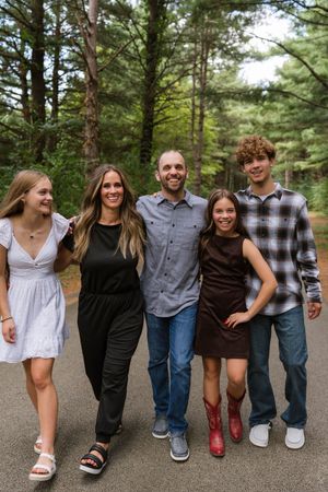 A smiling family of five poses together on a tree-lined path, dressed in casual outfits for an outdoor portrait.
