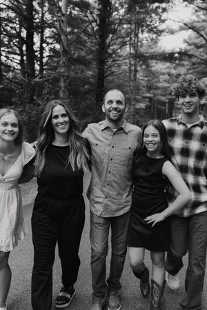 Black and white photo of a smiling family of five posing together outdoors among tall trees.