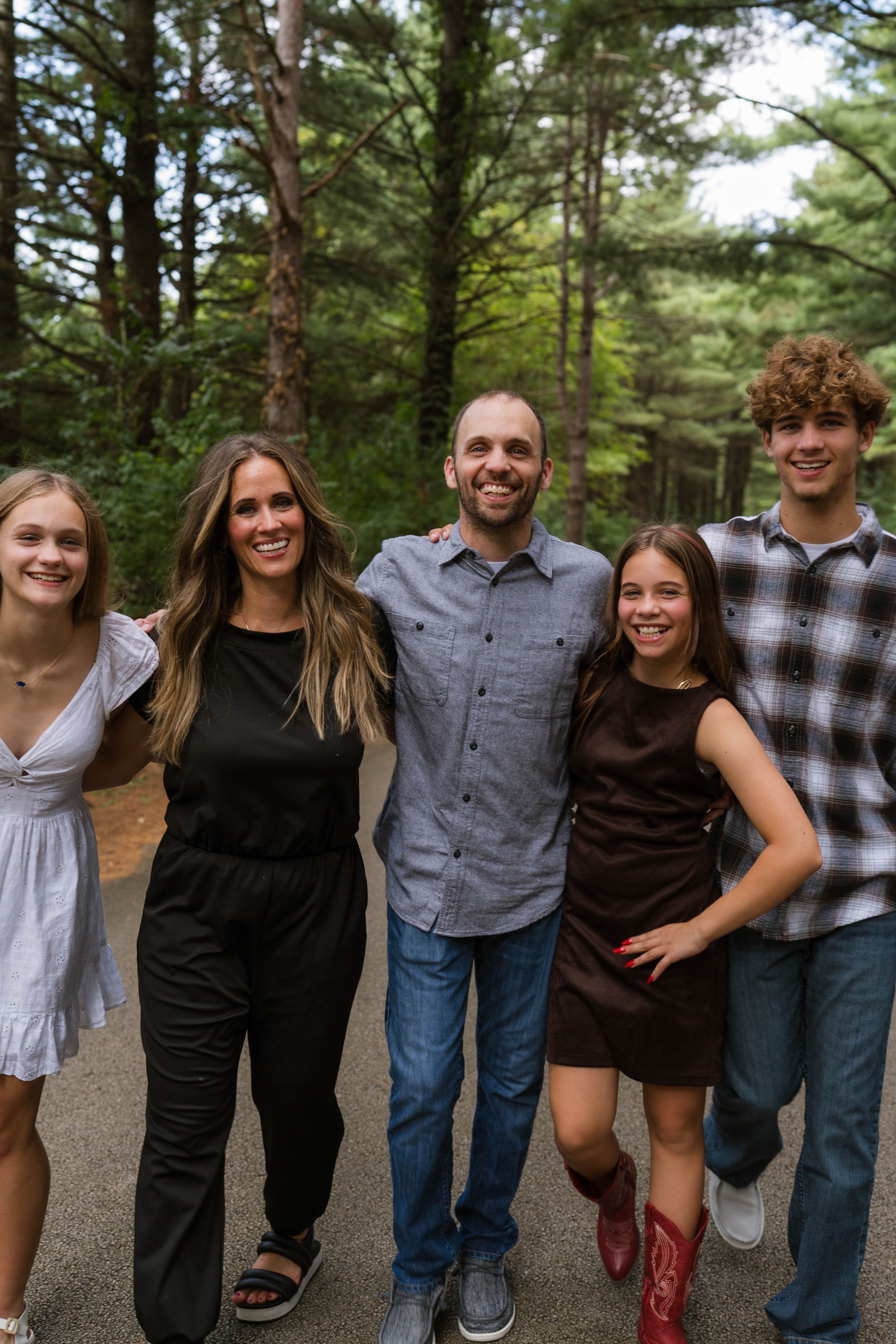 A smiling family of five poses together on a forest path surrounded by tall green trees.