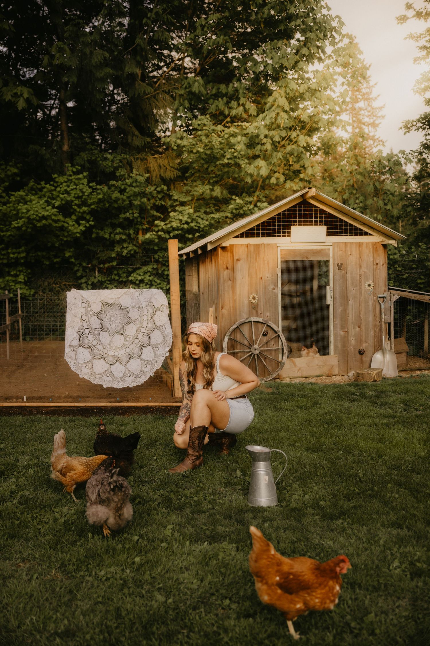 Woman in cowboy hat crouching near chickens beside rustic wooden chicken coop with lace decor in lush green backyard.