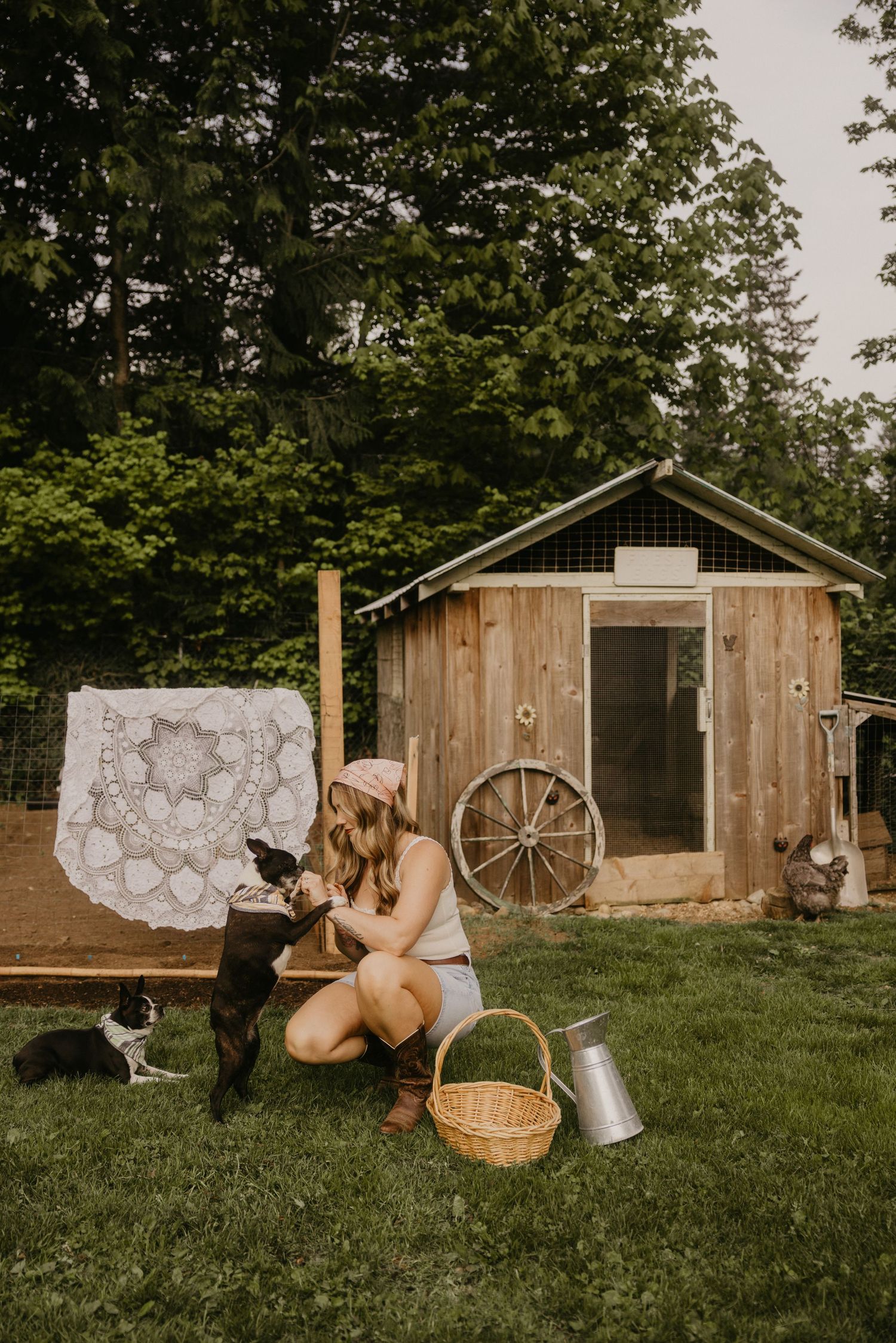 Woman kneeling on grass with dogs near rustic wooden shed, wicker basket, and watering can in a charming farmyard setting.