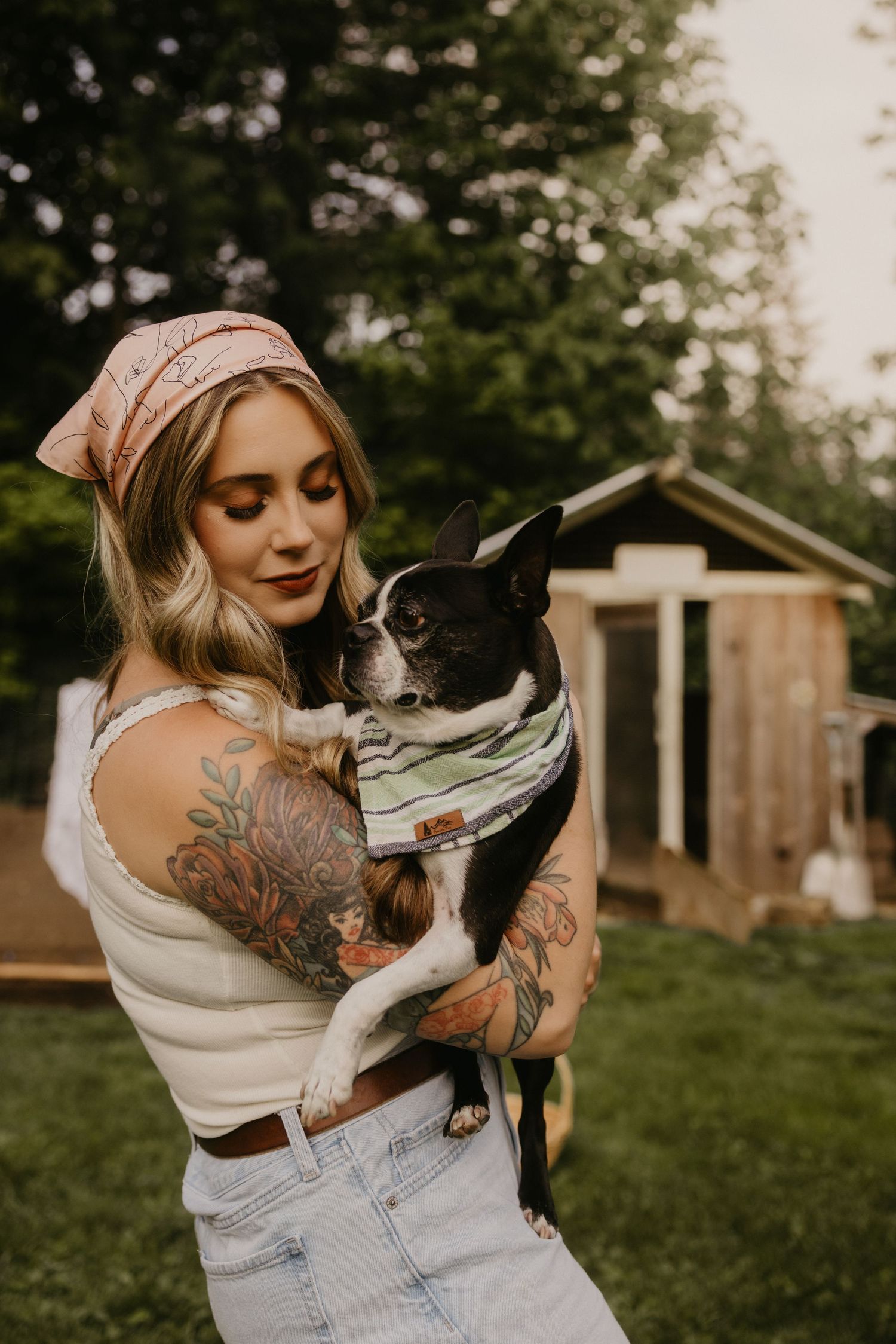 Tattooed woman holding a Boston Terrier dog wearing a striped bandana outdoors near a wooden shed.