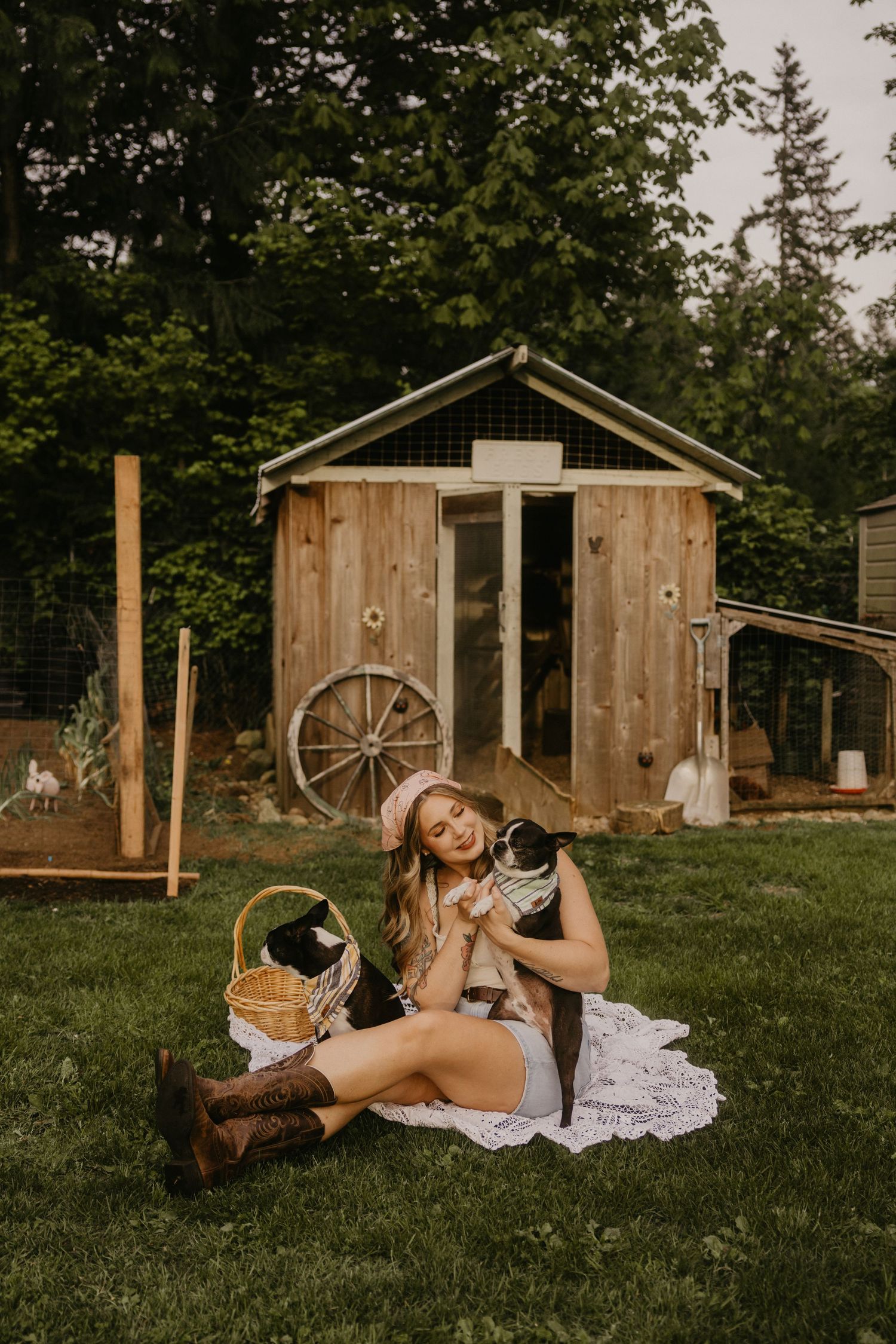 Woman in pink hat and boots sits on blanket holding a cat near a rustic wooden shed in a green backyard.