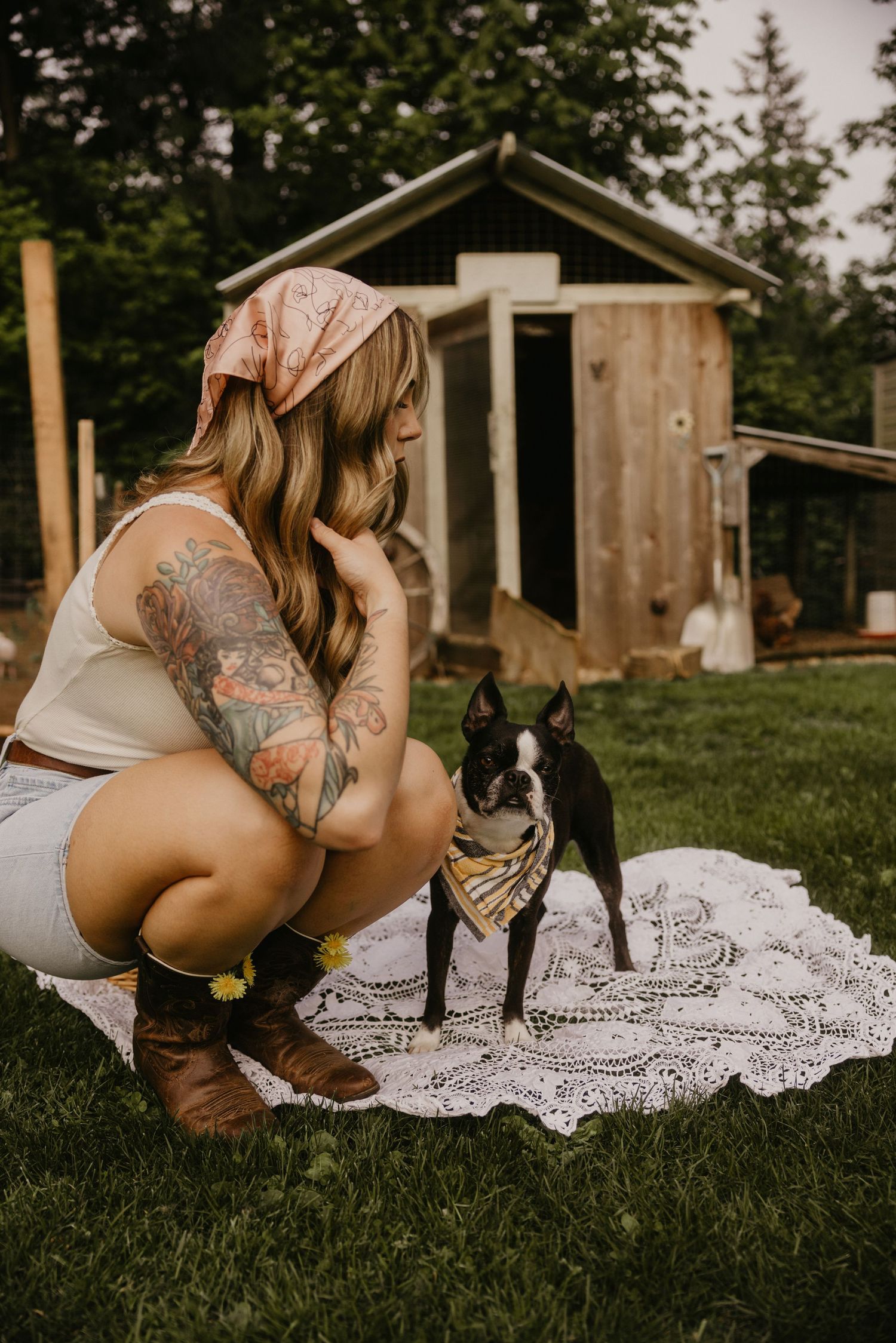Tattooed blonde woman in bandana crouching with Boston Terrier wearing bandana on lace blanket outdoors.