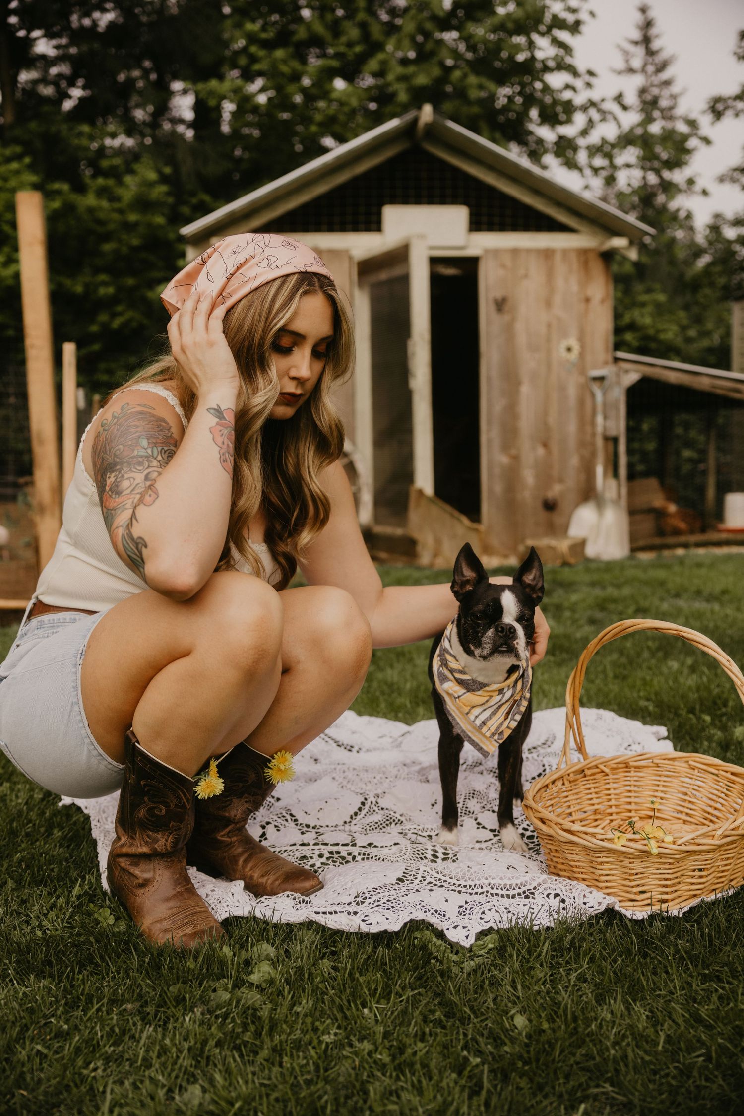 Woman in cowboy boots crouches beside a Boston Terrier wearing a bandana on a lace blanket in a rustic backyard.
