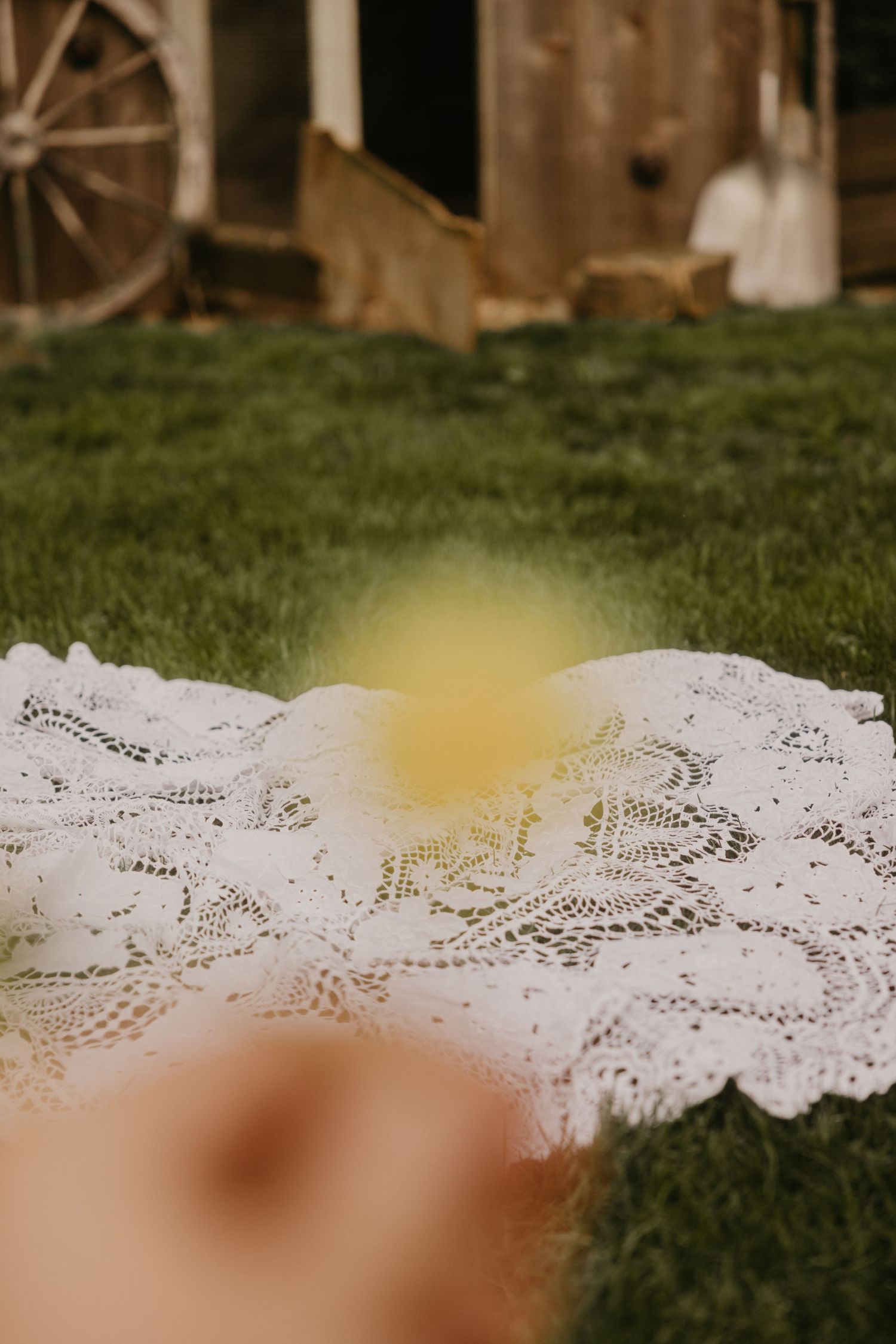 White lace blanket spread on green grass near a rustic wooden barn with an antique wagon wheel.