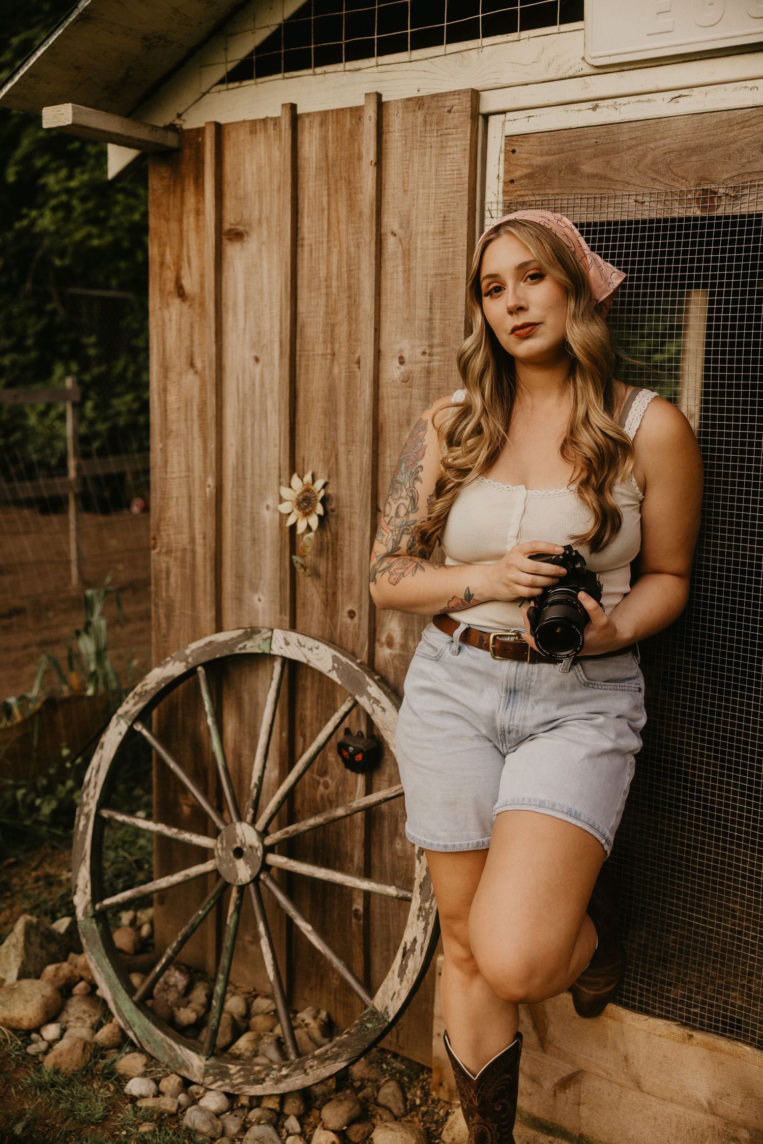 Young woman in denim shorts and cowboy boots holding a camera leaning against a rustic wooden fence.