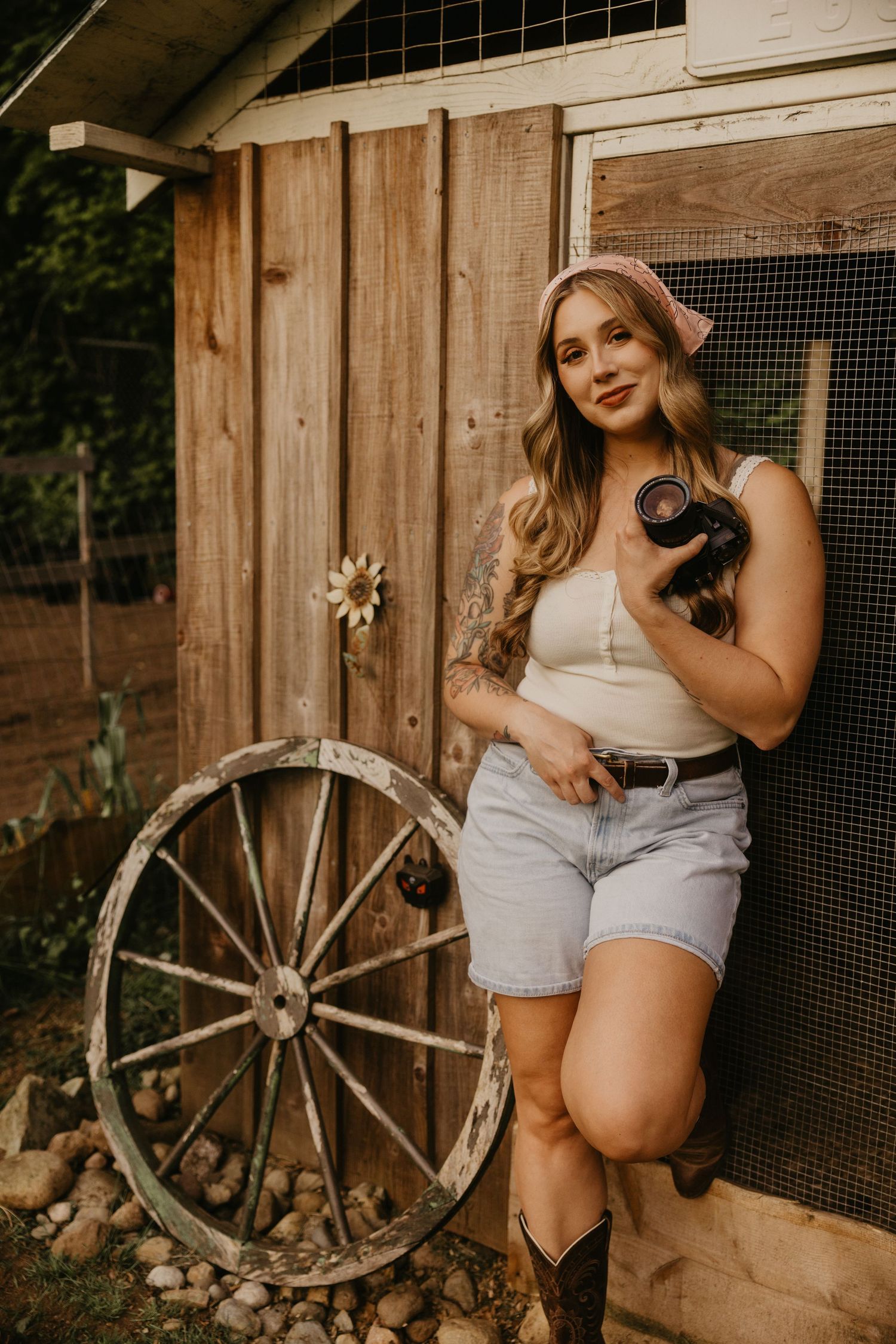 Young woman in denim shorts and cowboy boots holding a black chicken beside a rustic wooden barn with a vintage wagon wheel.