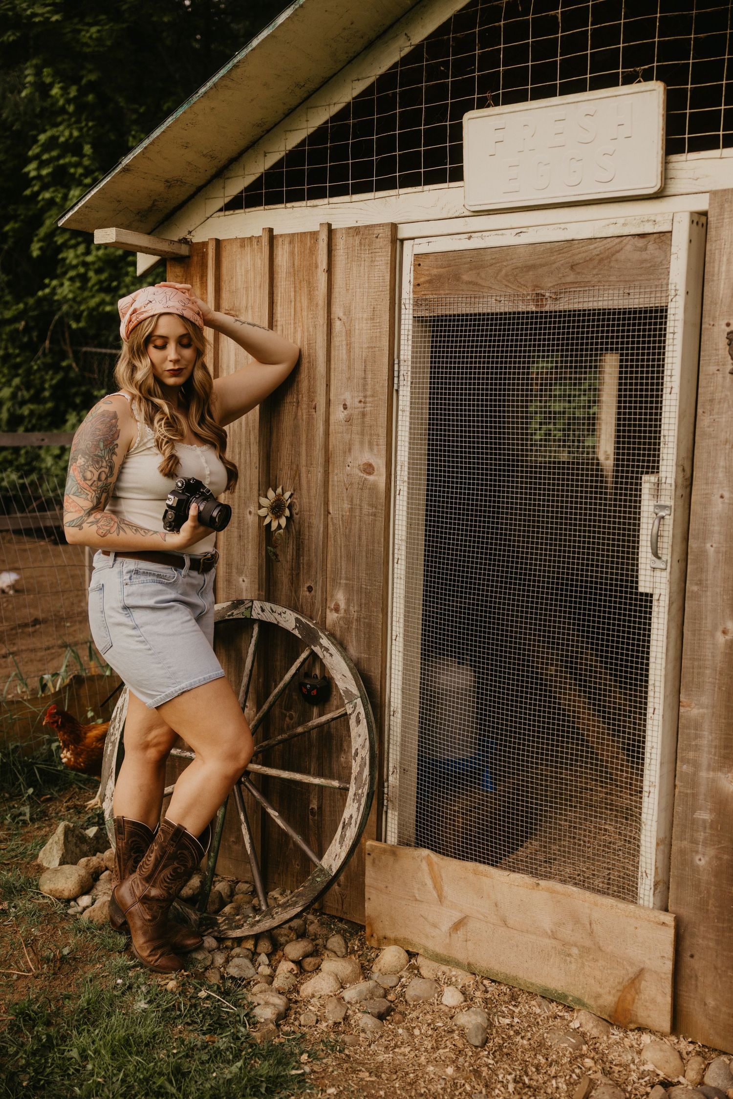 Tattooed blonde woman in bandana and cowboy boots posing by rustic wooden chicken coop with vintage wagon wheel.