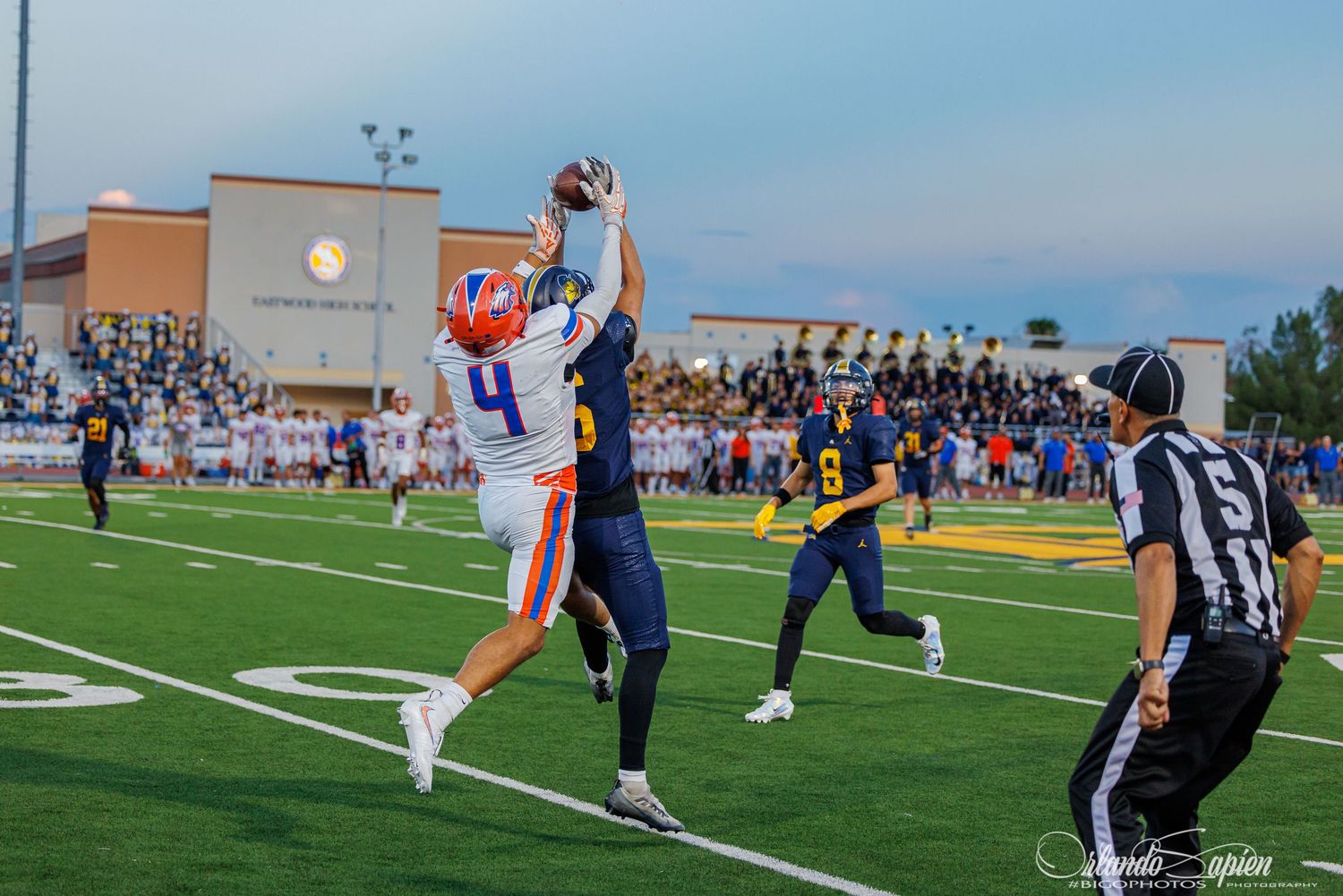 High school football player #4 leaps to catch a pass during a night game while a defender attempts to block.