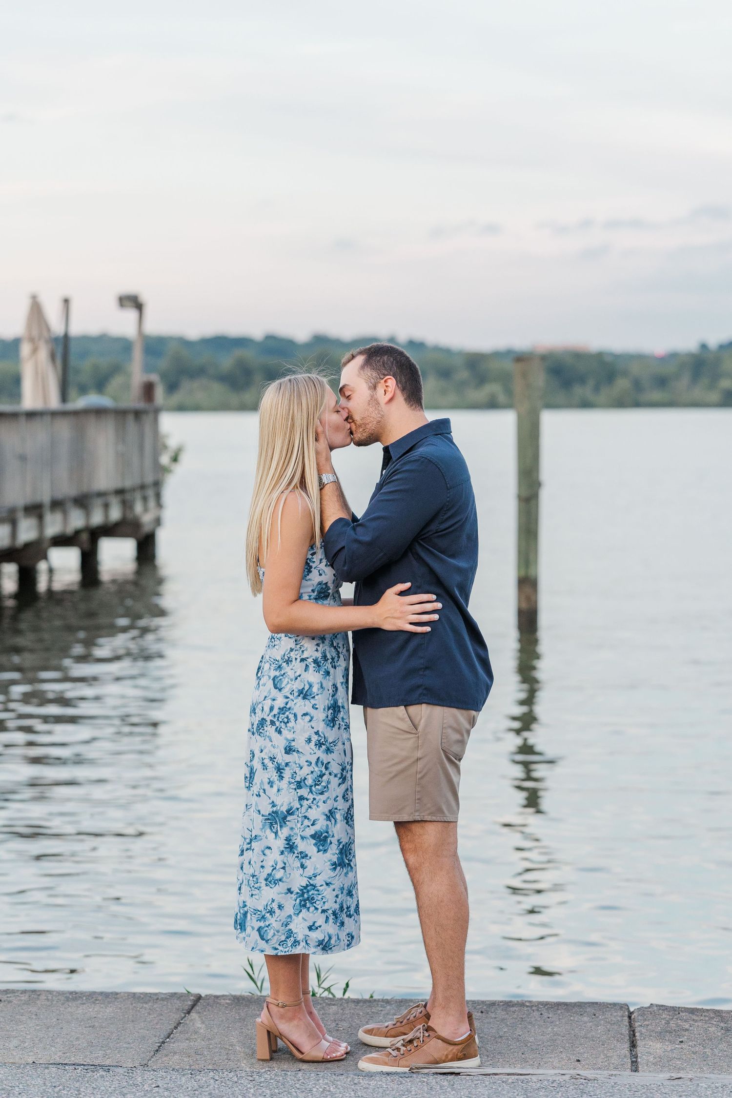 Couple sharing a romantic kiss on a waterfront dock, woman in floral dress, man in navy blazer and khaki shorts.