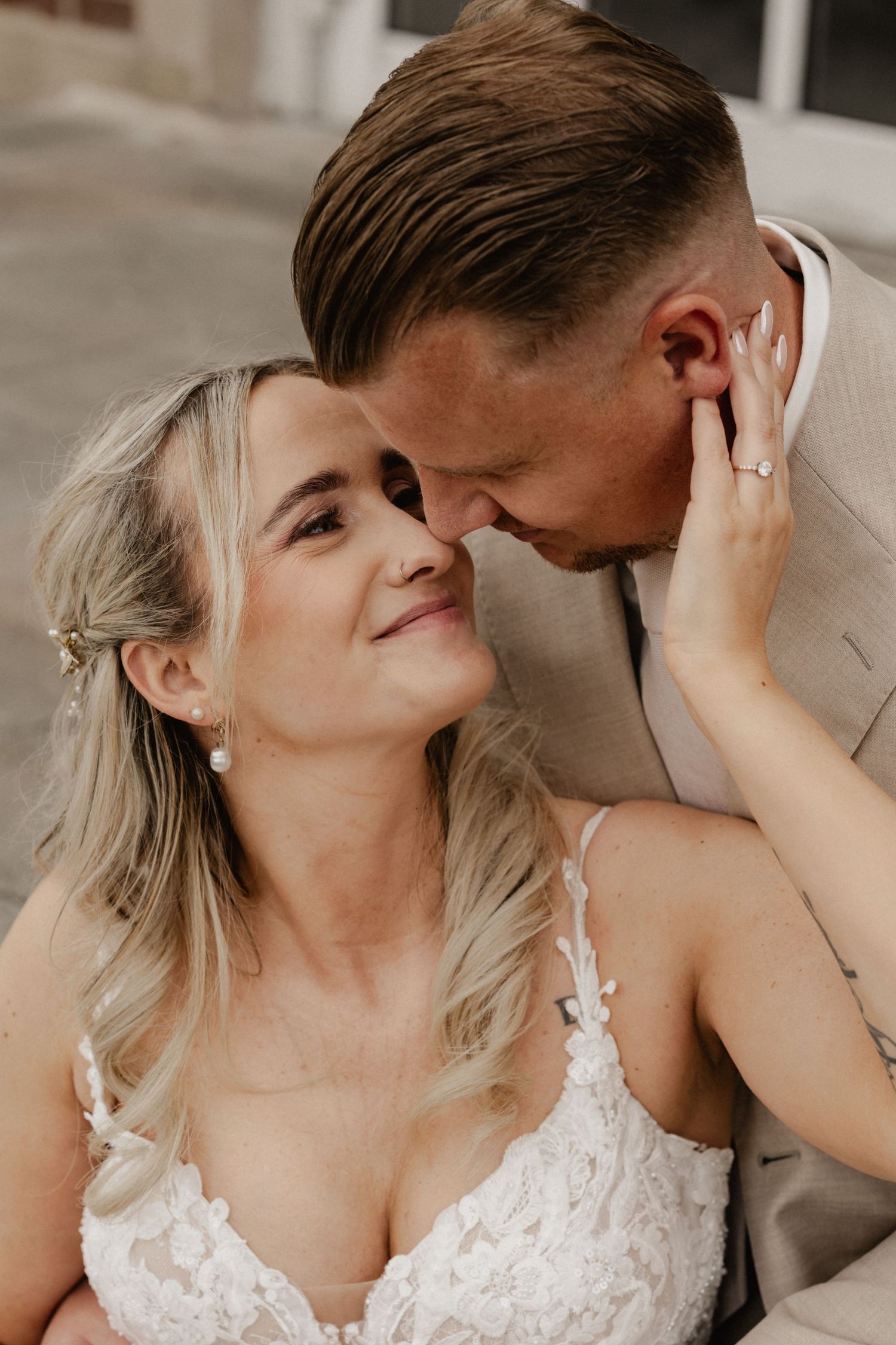 A bride in lace wedding dress smiles as groom in beige suit leans in for a kiss during romantic wedding portrait.