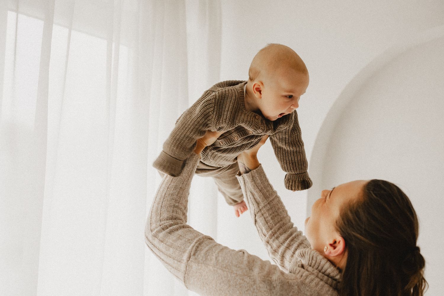 A joyful mother lifts her smiling baby in the air near a bright white curtained window.