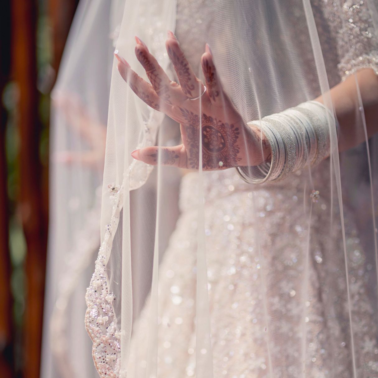 Bride's hand adorned with mehndi and bangles holding a beaded wedding veil over a sparkly white bridal gown.