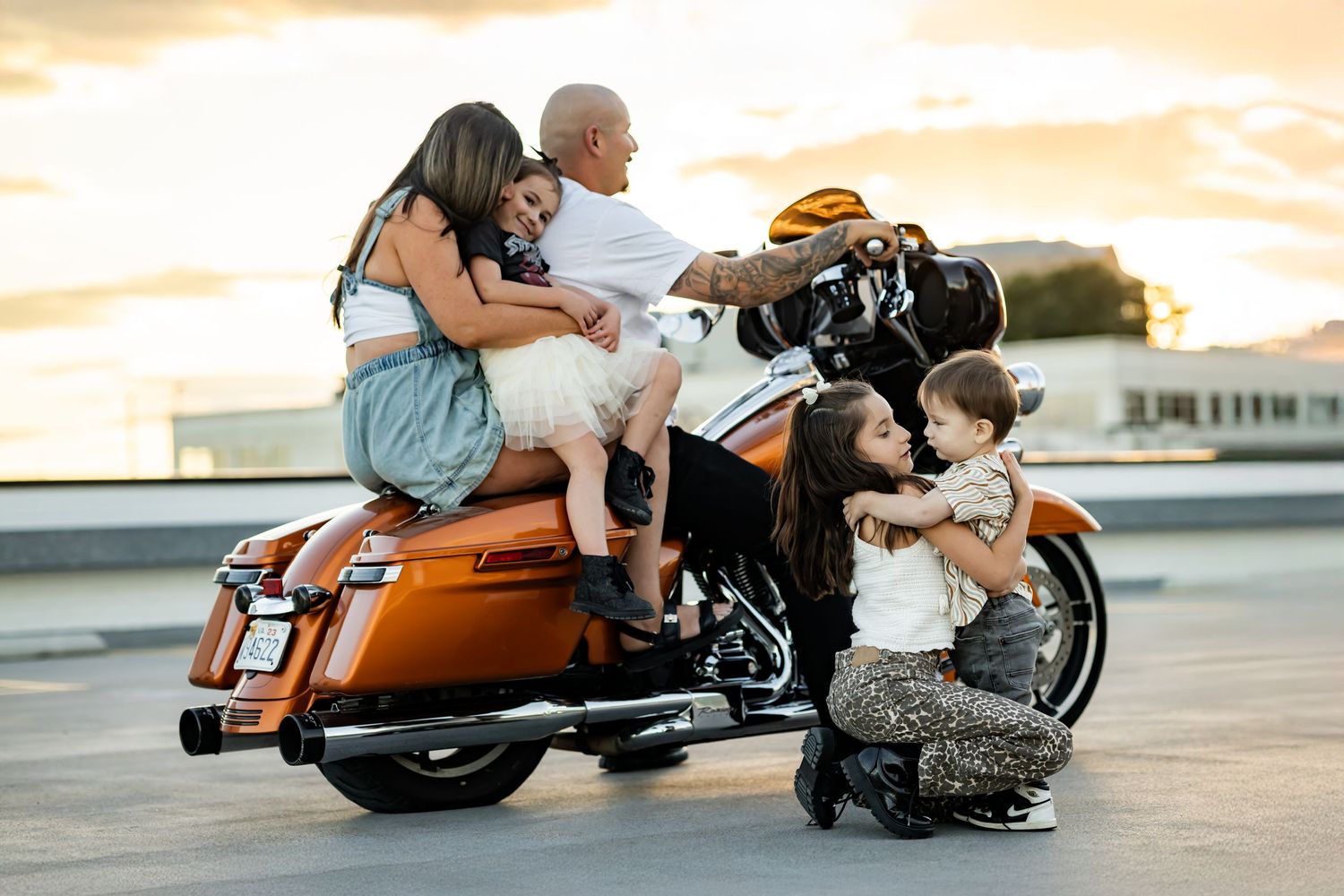Family posing together on an orange Harley-Davidson motorcycle at sunset on a rooftop parking lot.