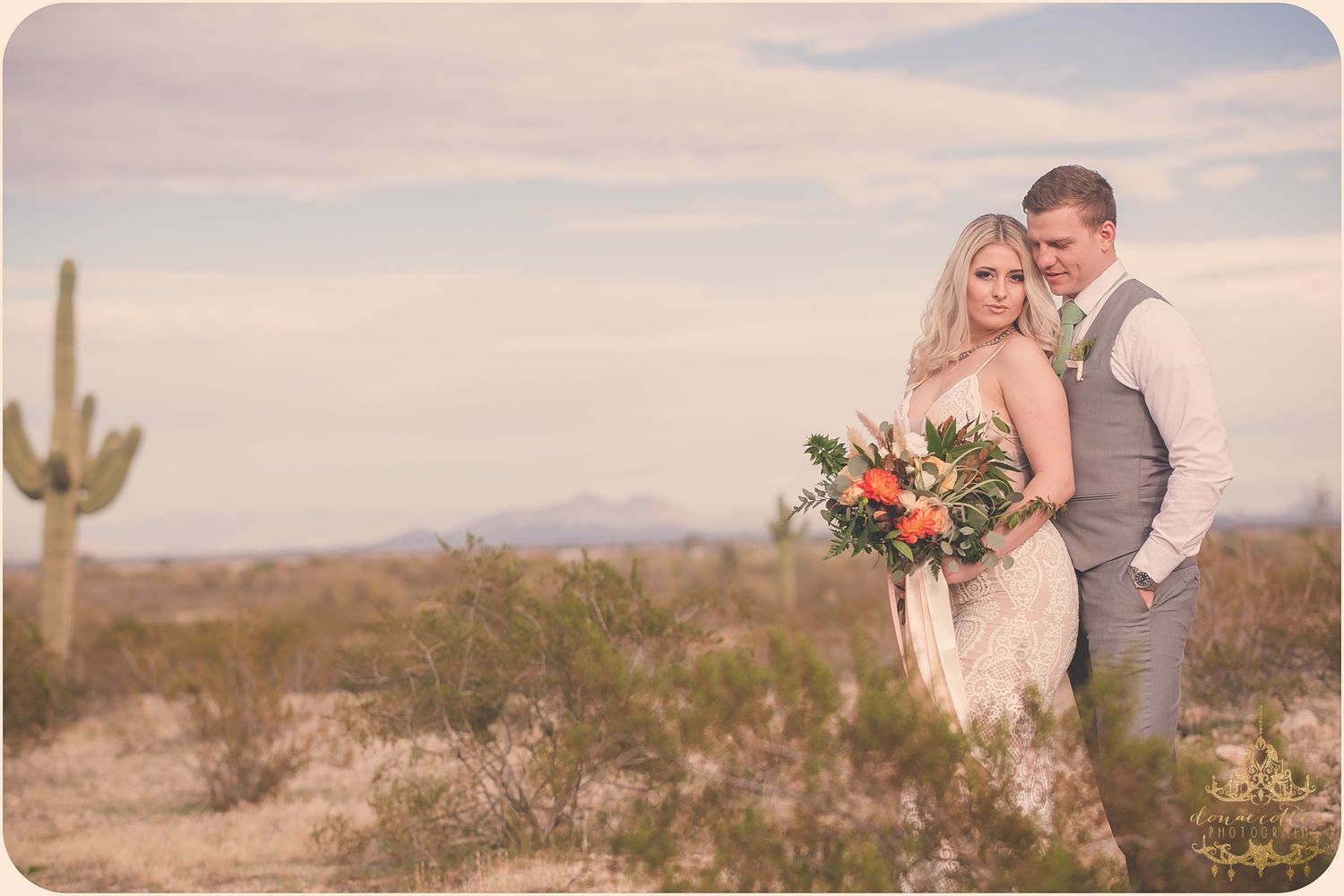 Bride and groom embrace in Arizona desert with saguaro cactus, bride holding vibrant bouquet, mountains in background.