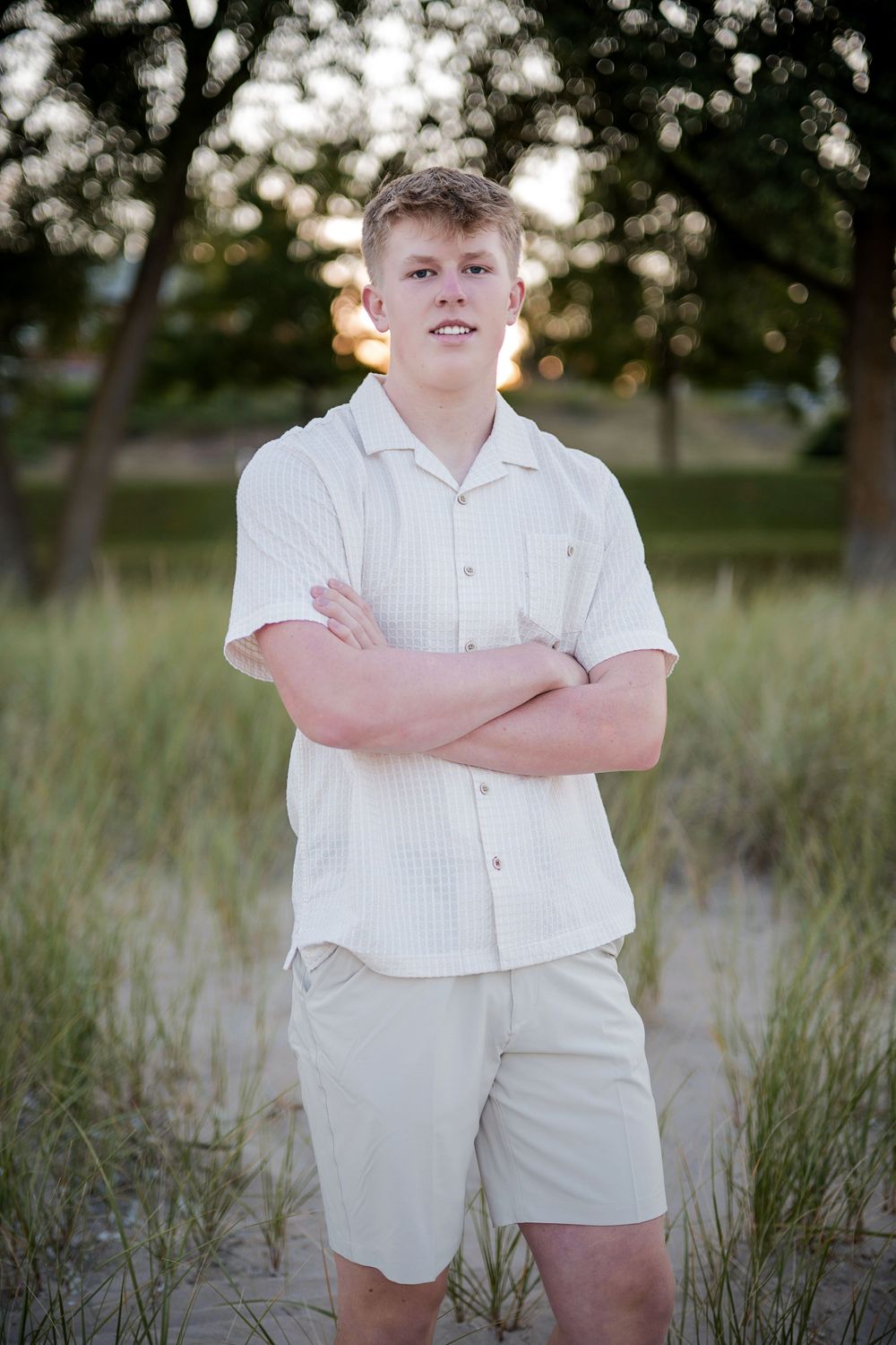 Young man in white shirt and khaki shorts standing with arms crossed on sandy beach at sunset.