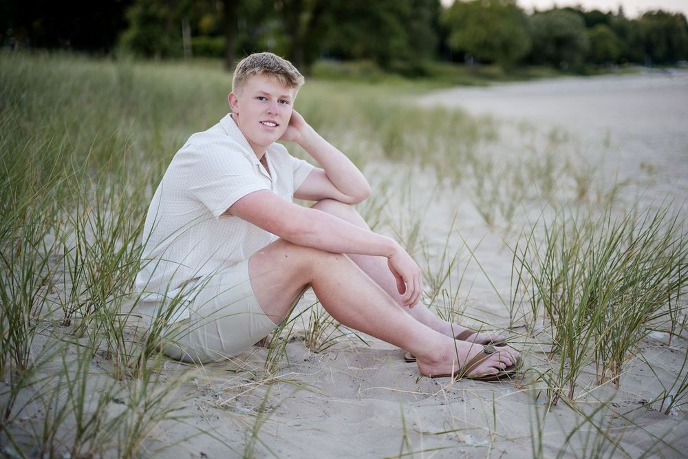 Young blonde teen boy sitting on sandy beach dunes with grass, wearing white shirt and sandals, smiling outdoors.