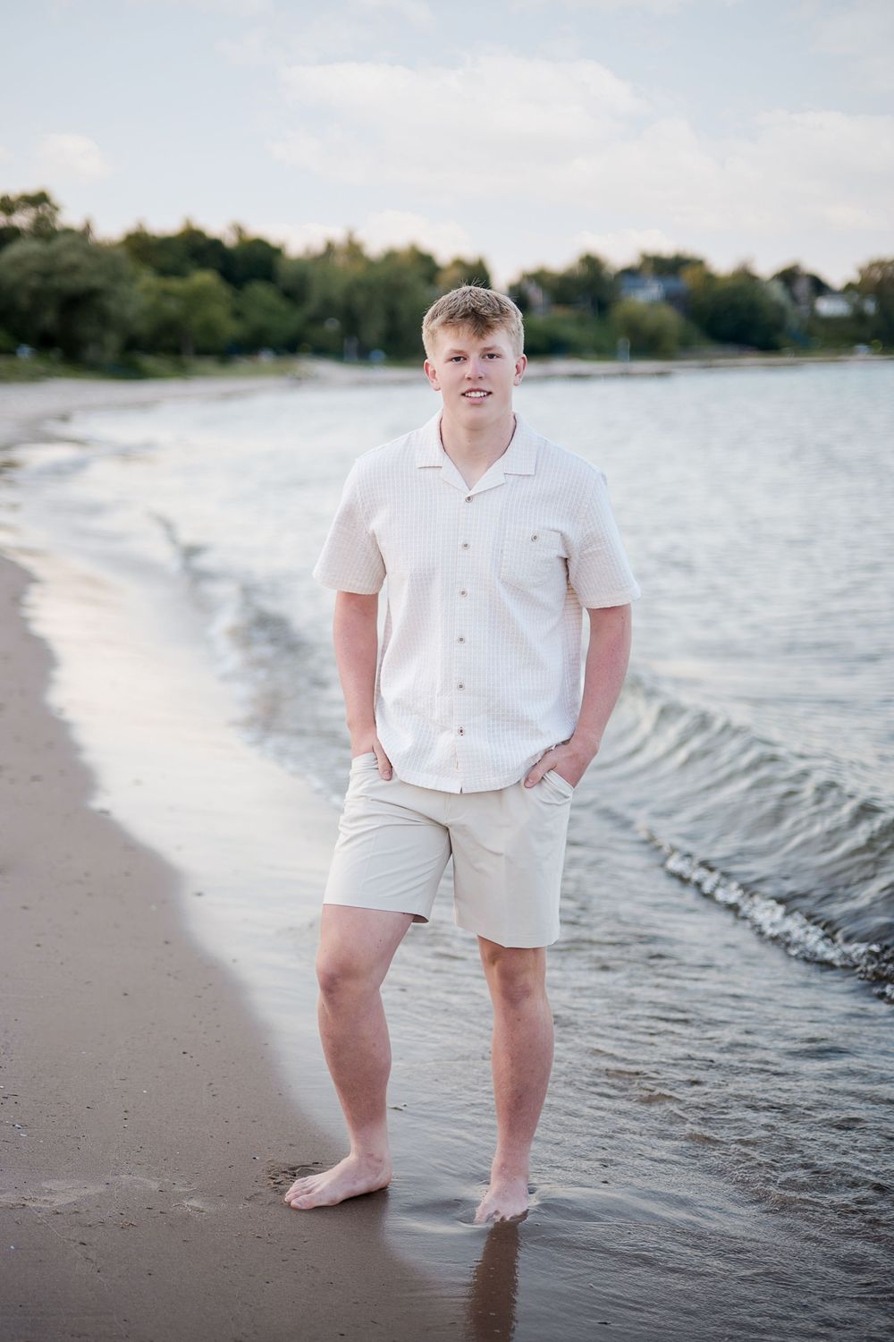 Young man in white shirt and khaki shorts standing barefoot on a sandy beach with gentle waves.