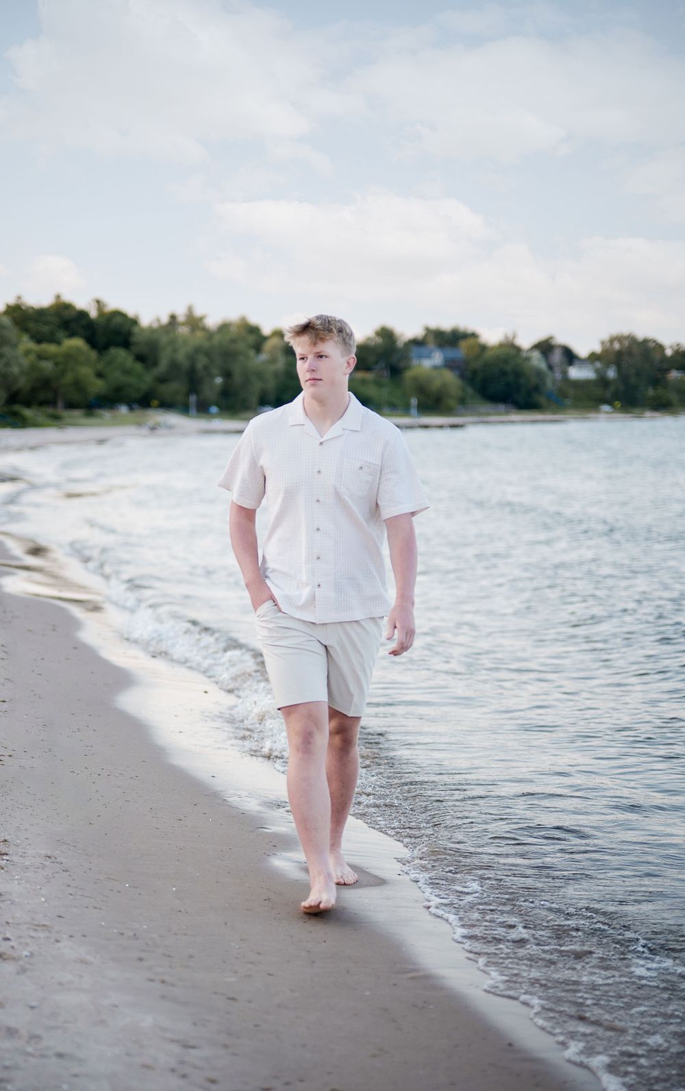 Young man in white shirt and khaki shorts walking barefoot along a sandy beach shoreline with calm water.