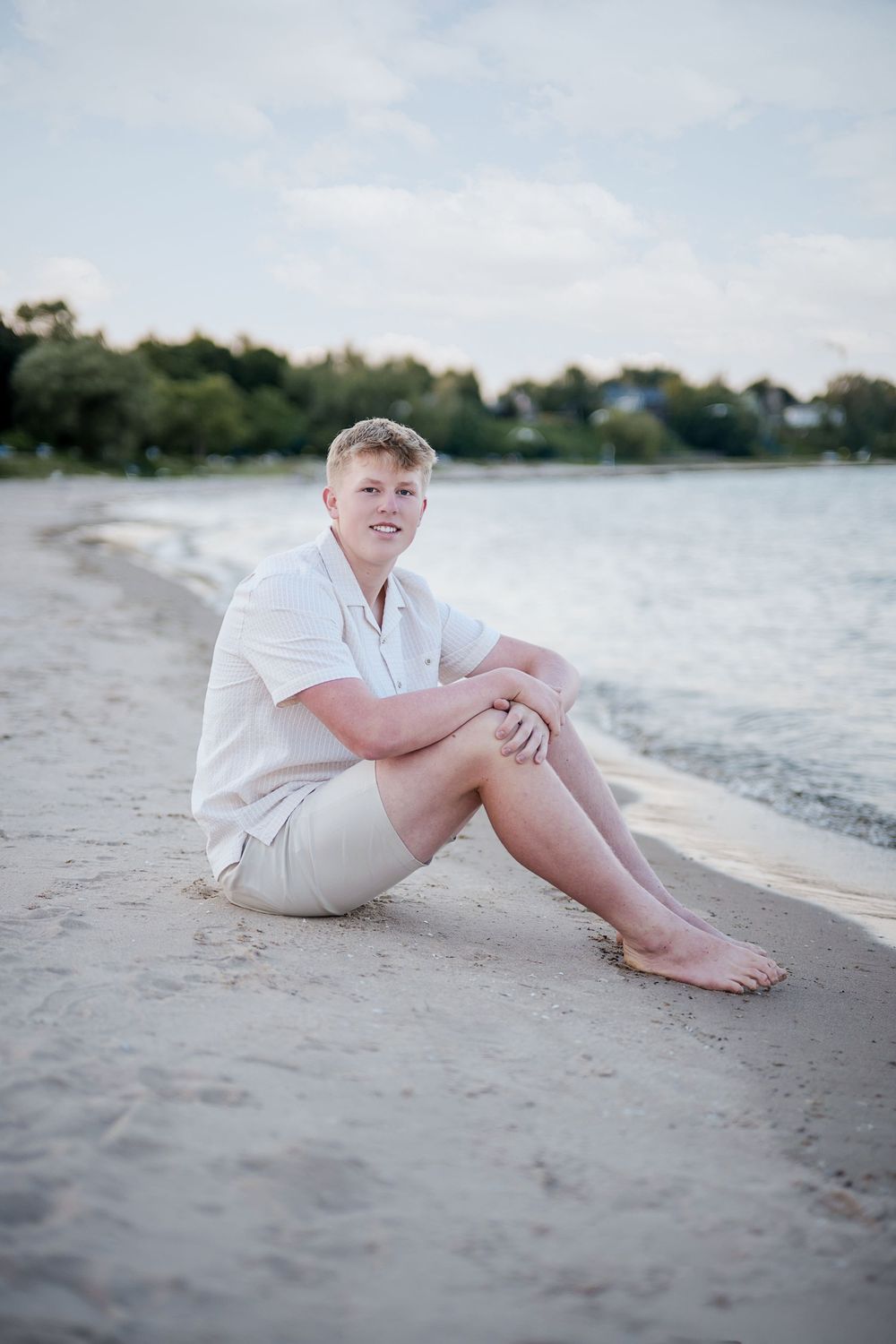 Young man in white shirt and khaki shorts sitting barefoot on sandy beach shore with calm water and trees behind.