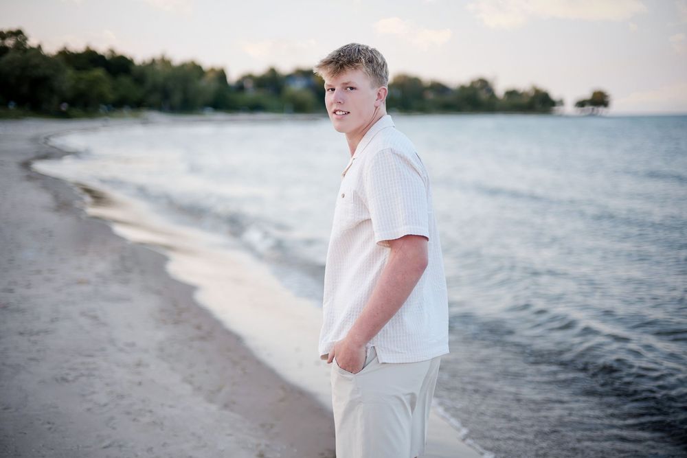 Young man in white shirt and khaki pants posing on a sandy beach shoreline at golden hour.