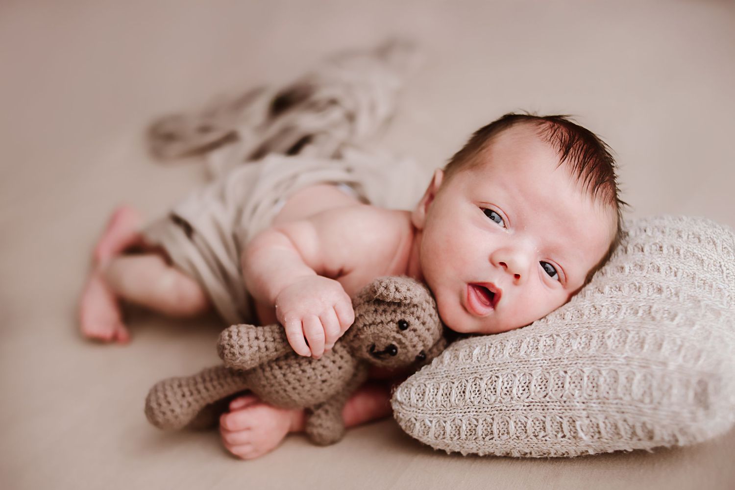 Newborn baby lying on a soft blanket holding a crocheted teddy bear, with a knit pillow in a neutral studio setting.
