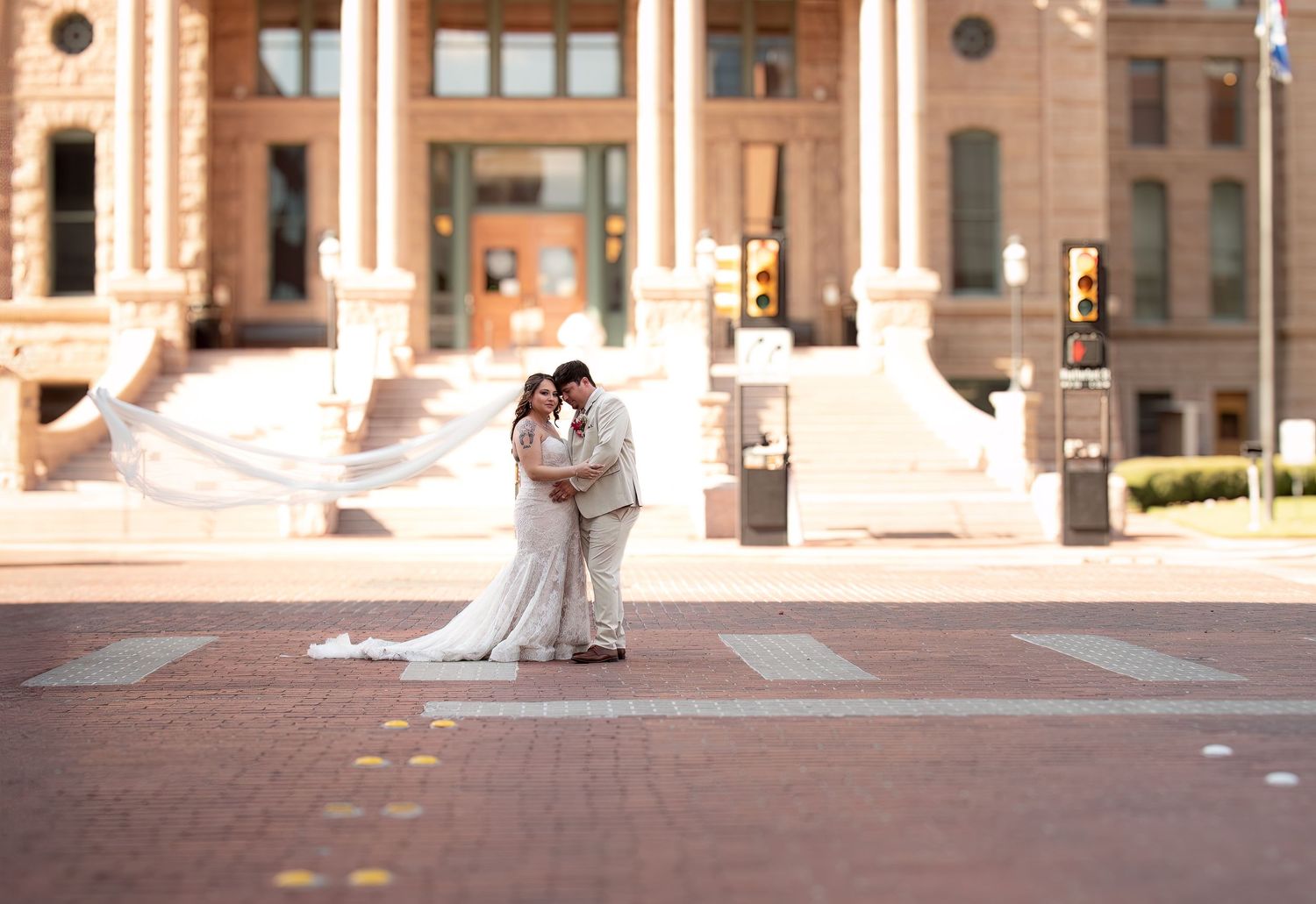 Bride and groom share a kiss at a crosswalk in front of a historic brick building, with veil flowing in the wind.