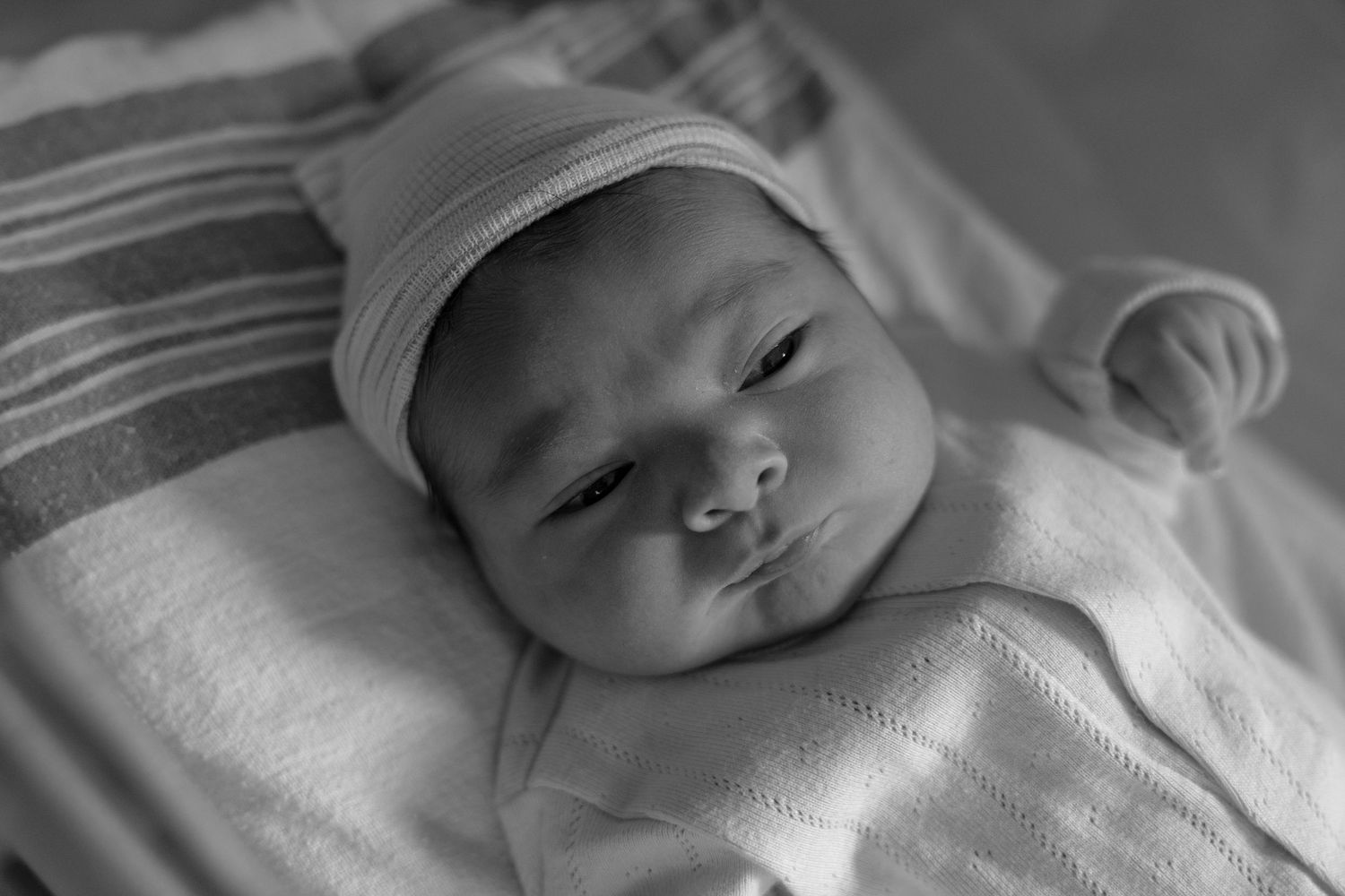 Black and white photo of a newborn baby wearing a white hat, lying on a striped blanket.