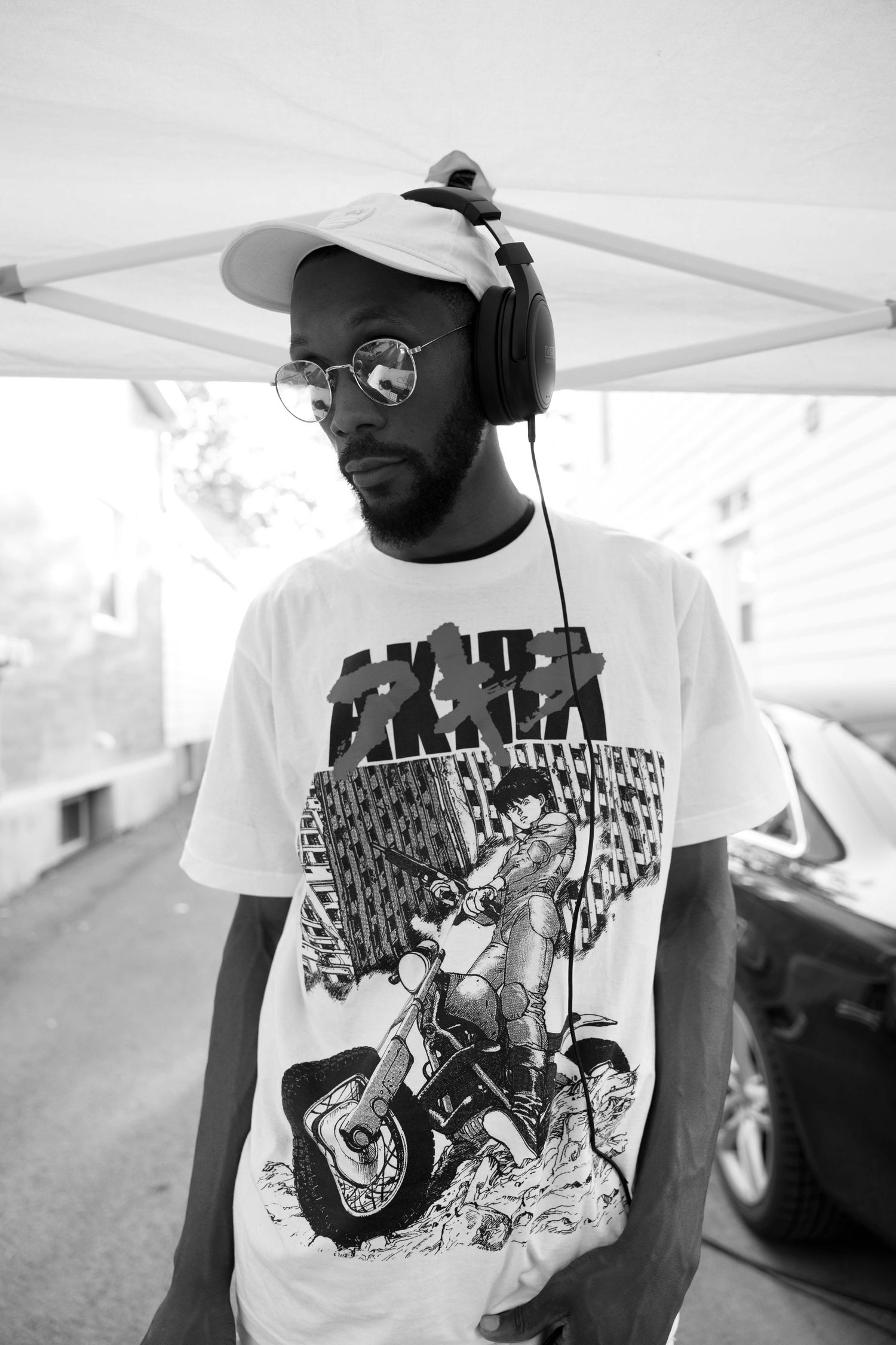Black and white photo of a young man wearing headphones, cap, glasses, and anime graphic tee outdoors.