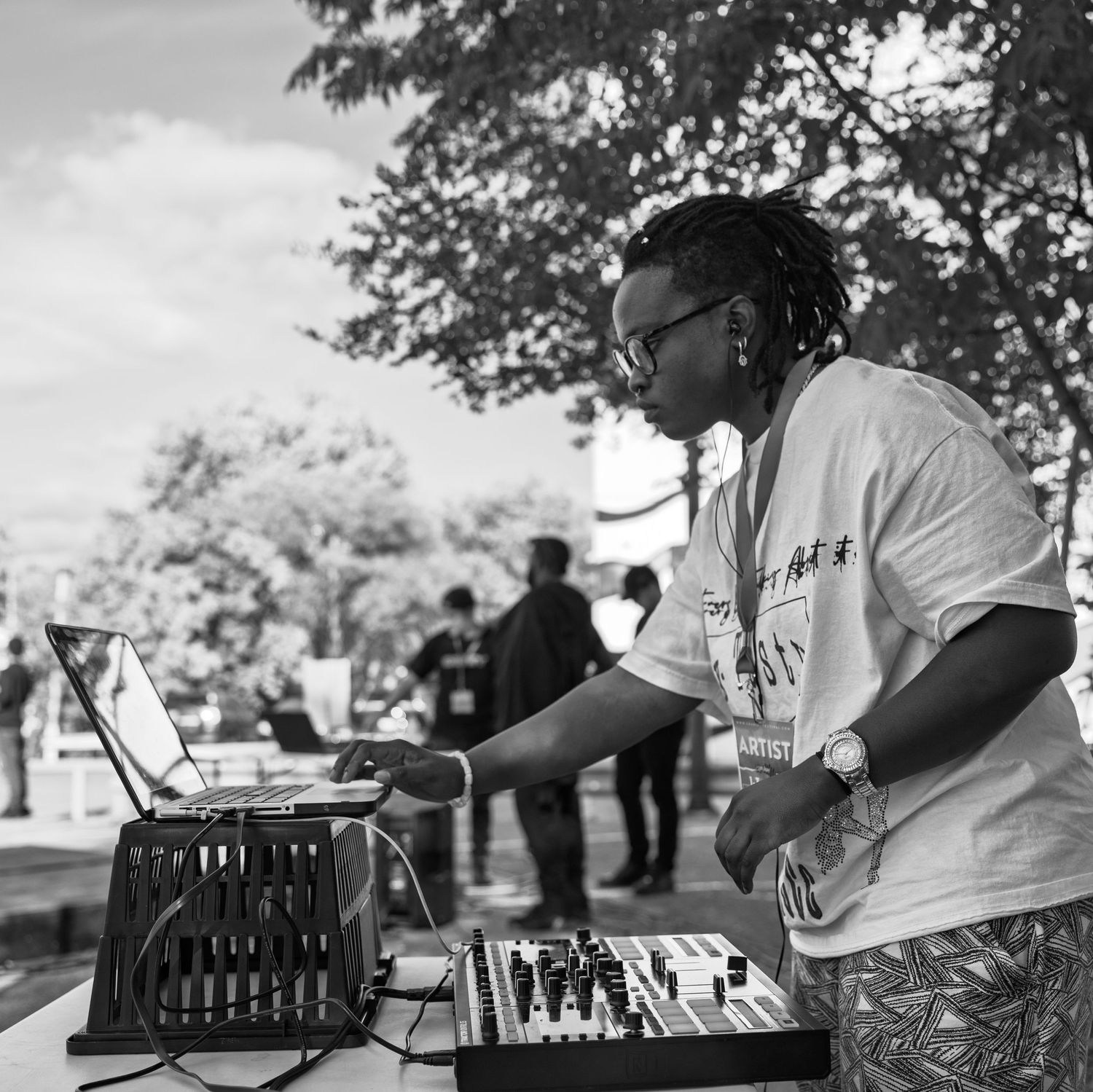 Black and white photo of a DJ mixing music outdoors at an event, operating a mixer and laptop under trees.