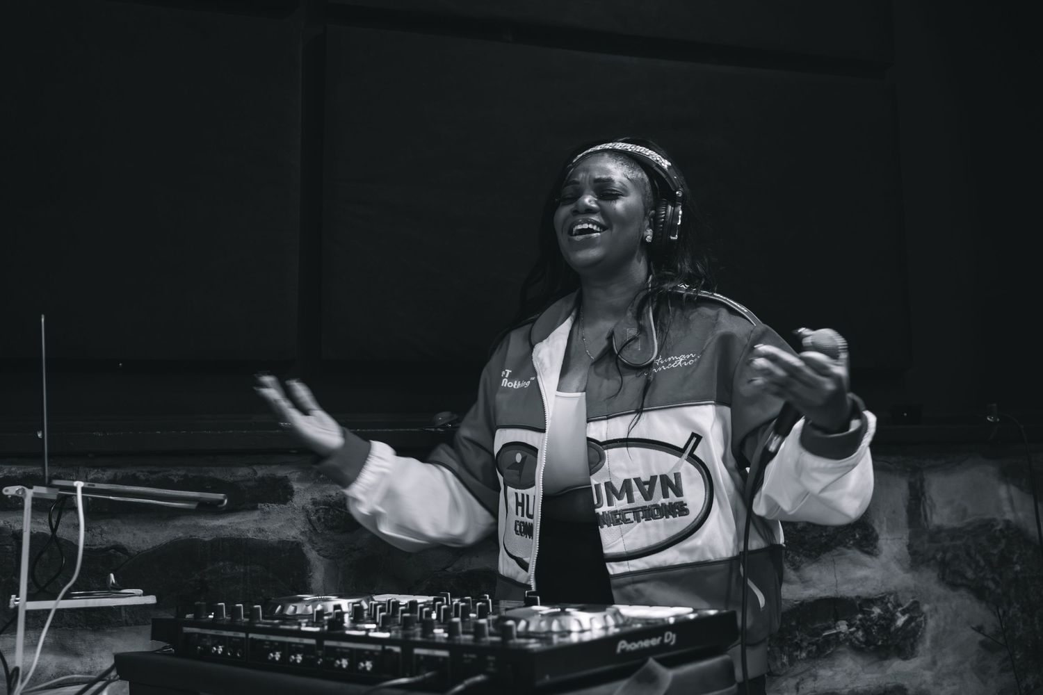 Female DJ performing at turntables in black and white photo, wearing jacket and headphones around neck.