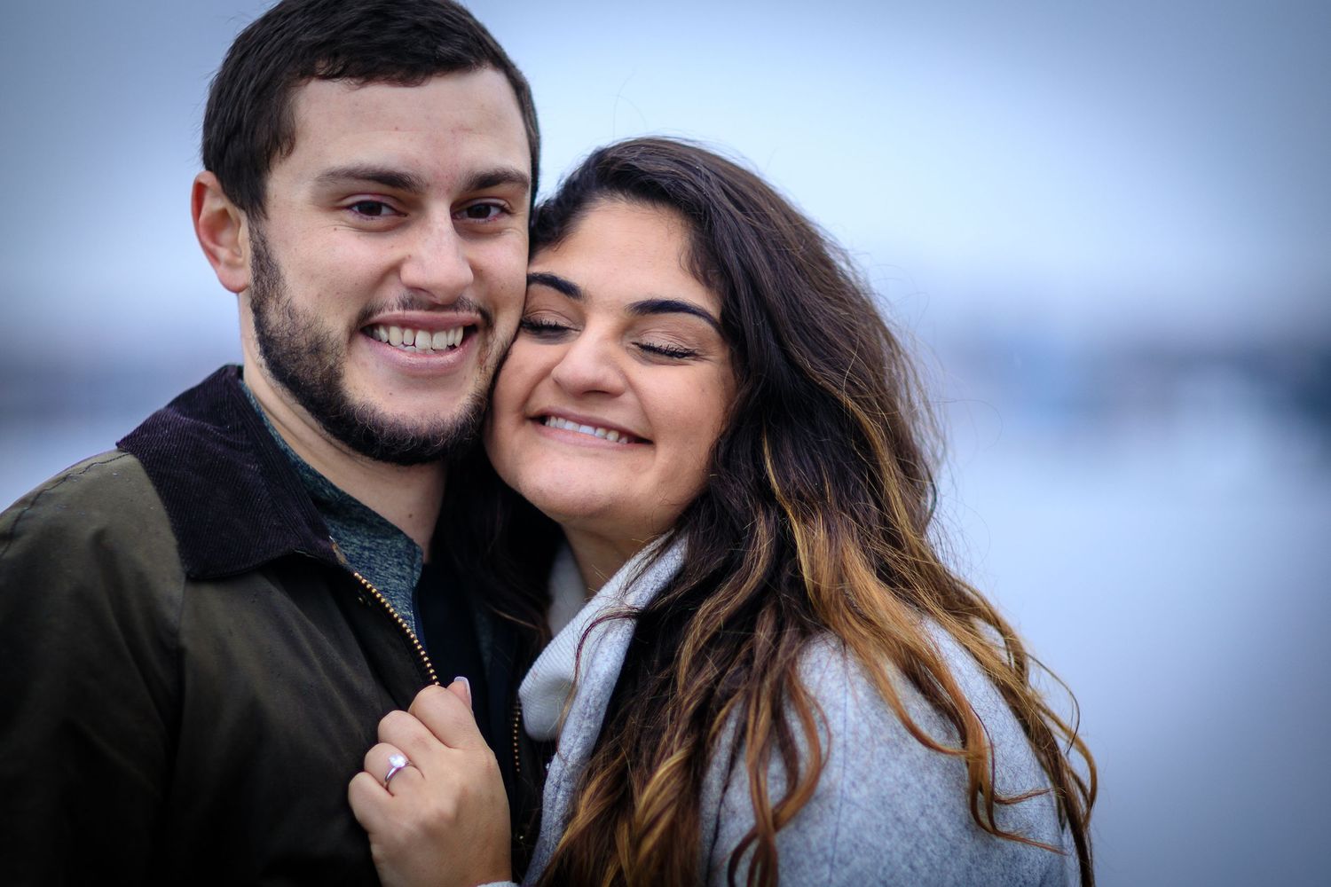 A smiling couple embraces outdoors in winter, woman with long brown hair leans on mans shoulder near waterfront.