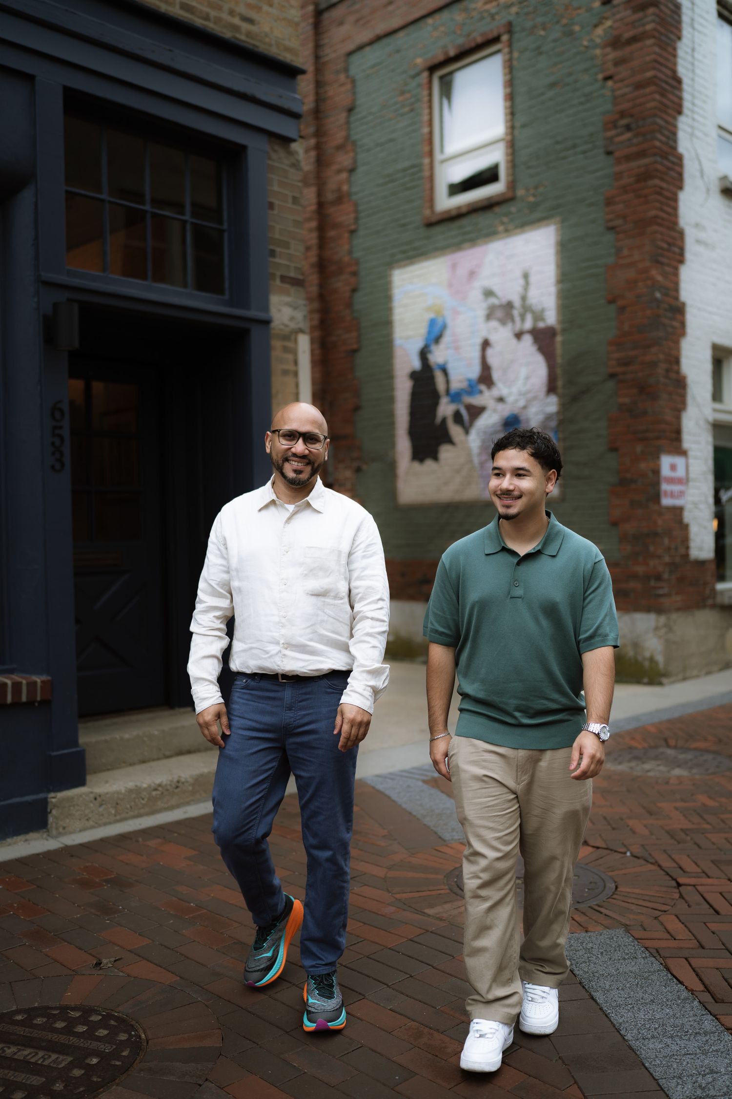 Two men walking together on a brick alley street past a colorful mural on a brick building.