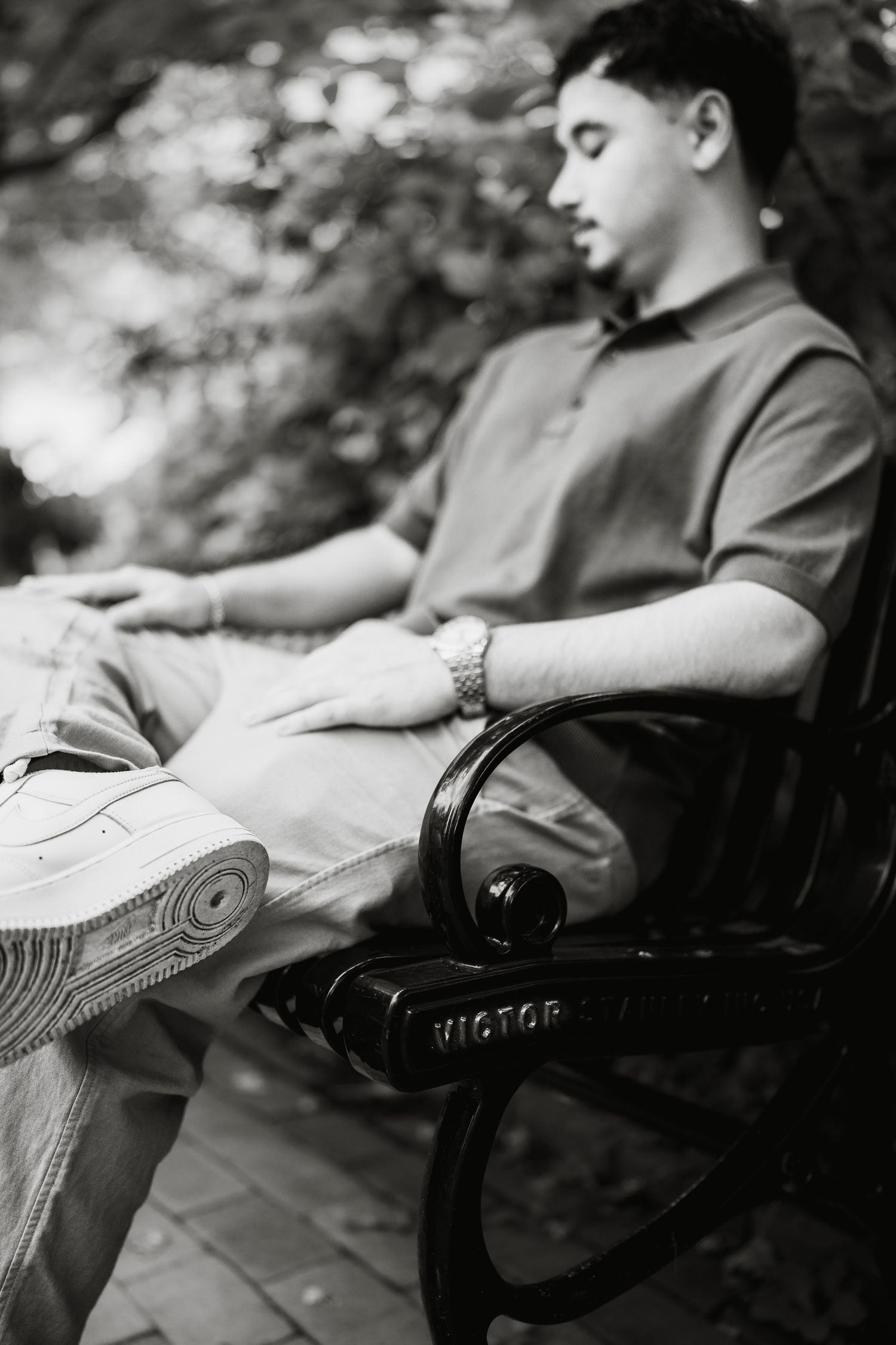 Young man relaxing on a Victor park bench outdoors, wearing a polo shirt and sneakers, in black and white photo.
