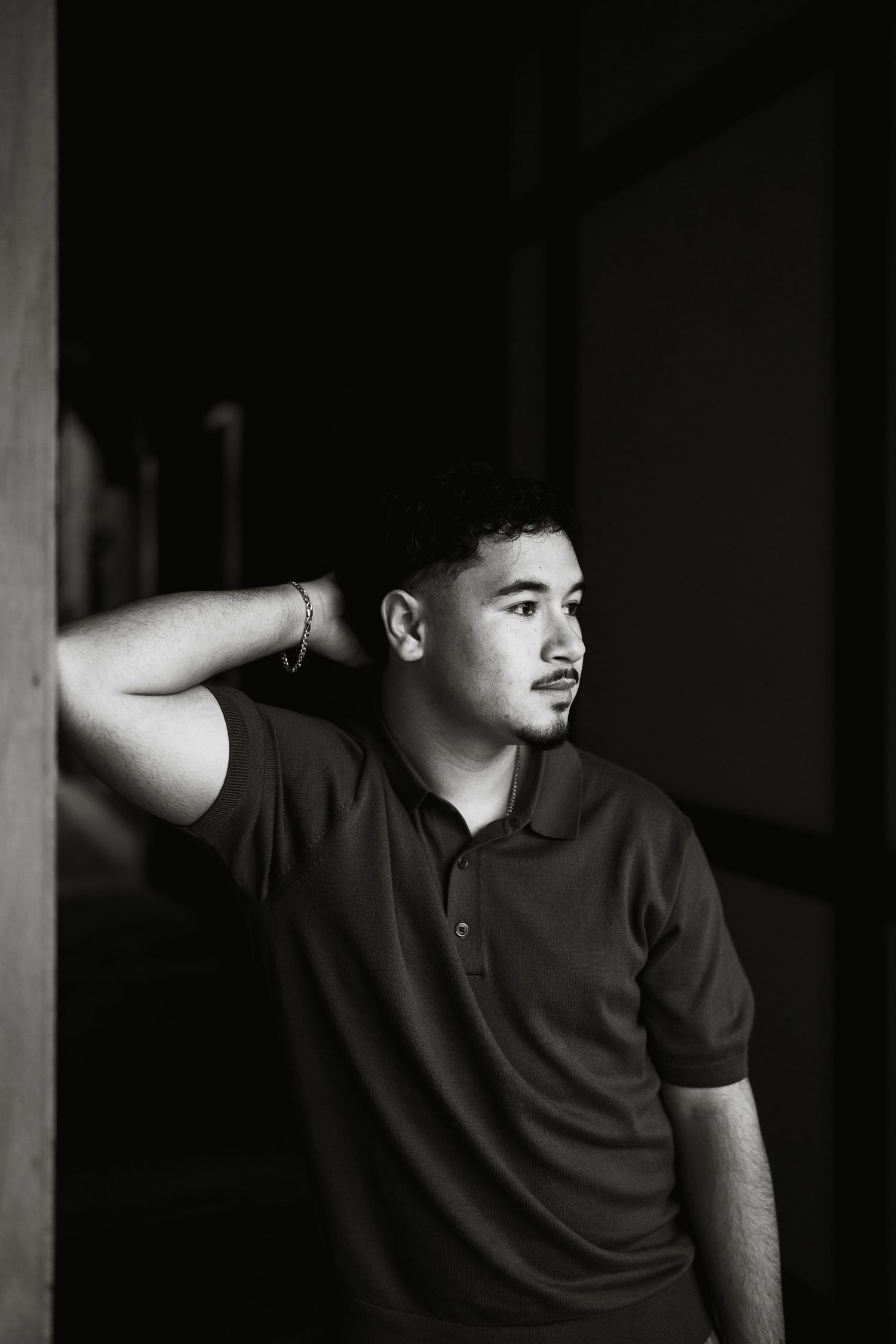 Black and white portrait of a young man in a polo shirt leaning against a wall with hand behind his neck.
