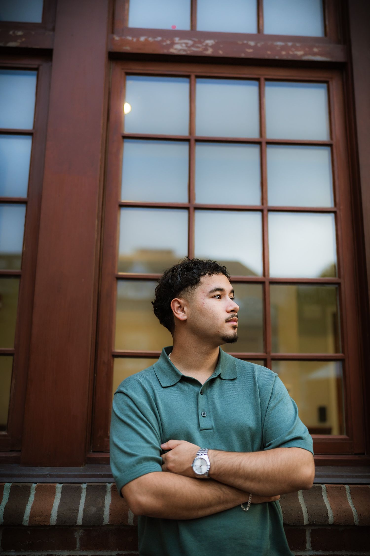 Young man in green polo with crossed arms stands thoughtfully before a large wooden framed window.