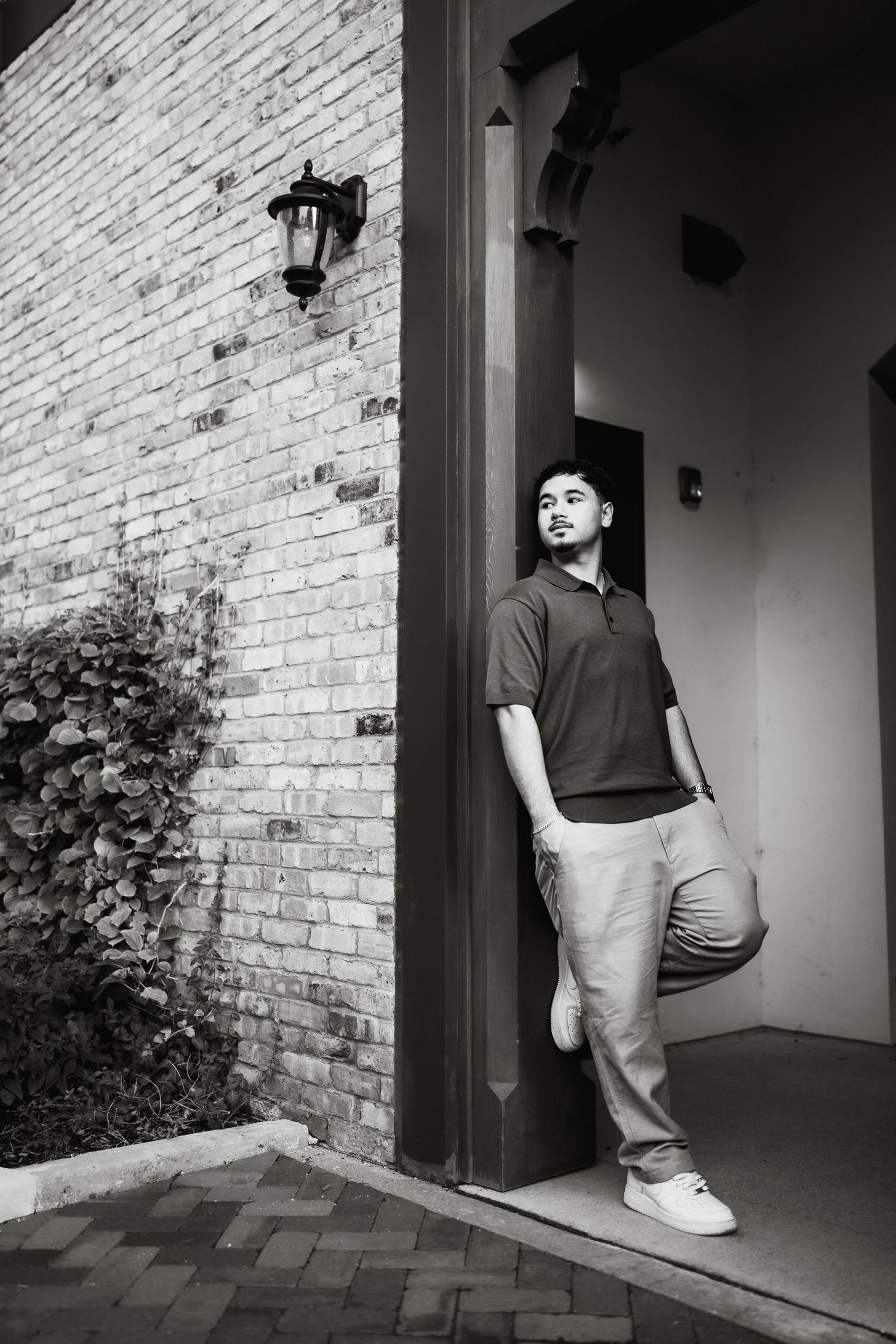 Black and white photo of a young man leaning against a brick building doorway, wearing a polo shirt and light pants.