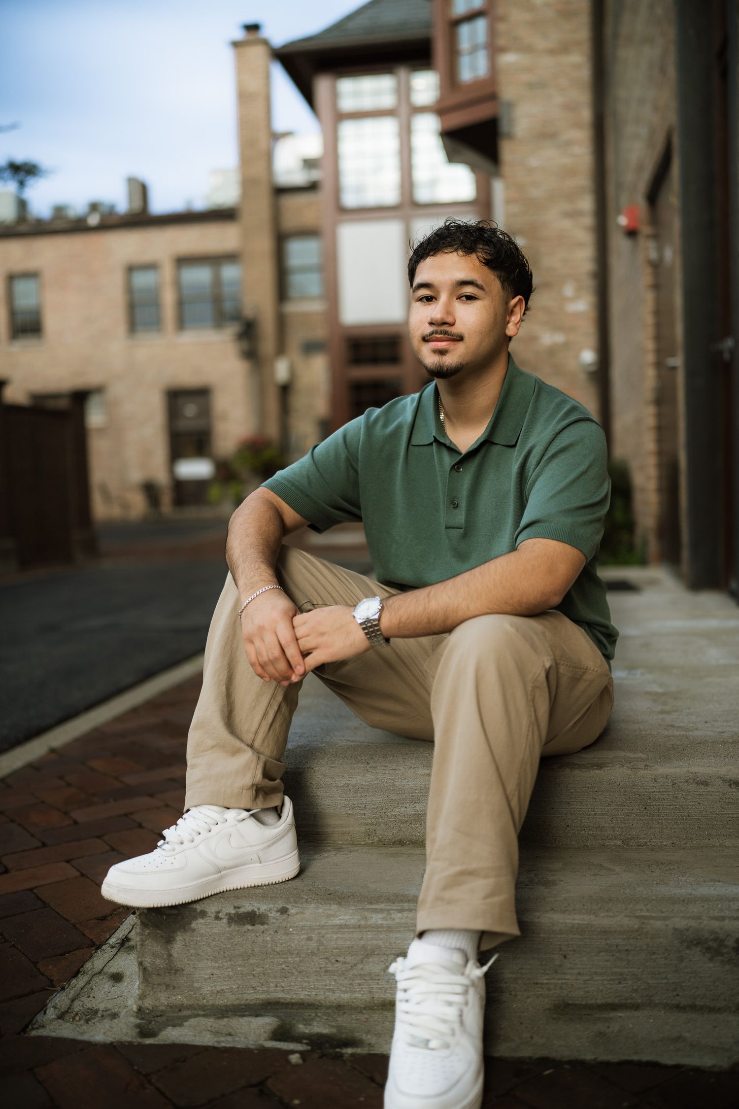 Young man in green polo and khaki pants sitting on steps outdoors near brick buildings wearing white sneakers.