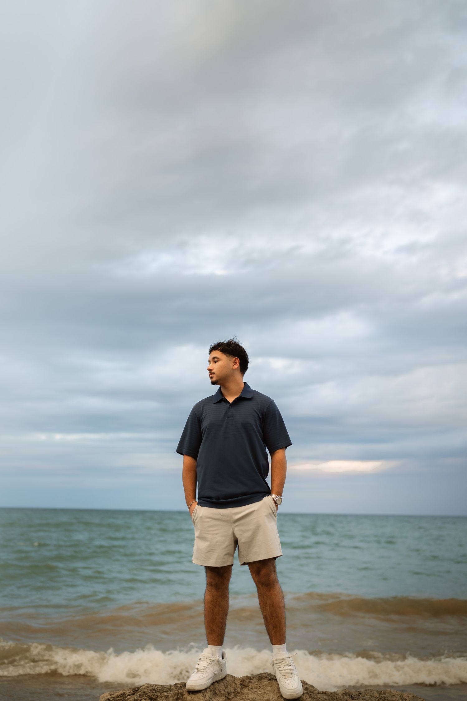 Young man in dark polo shirt and beige shorts standing on beach shore with cloudy sky and ocean in background.