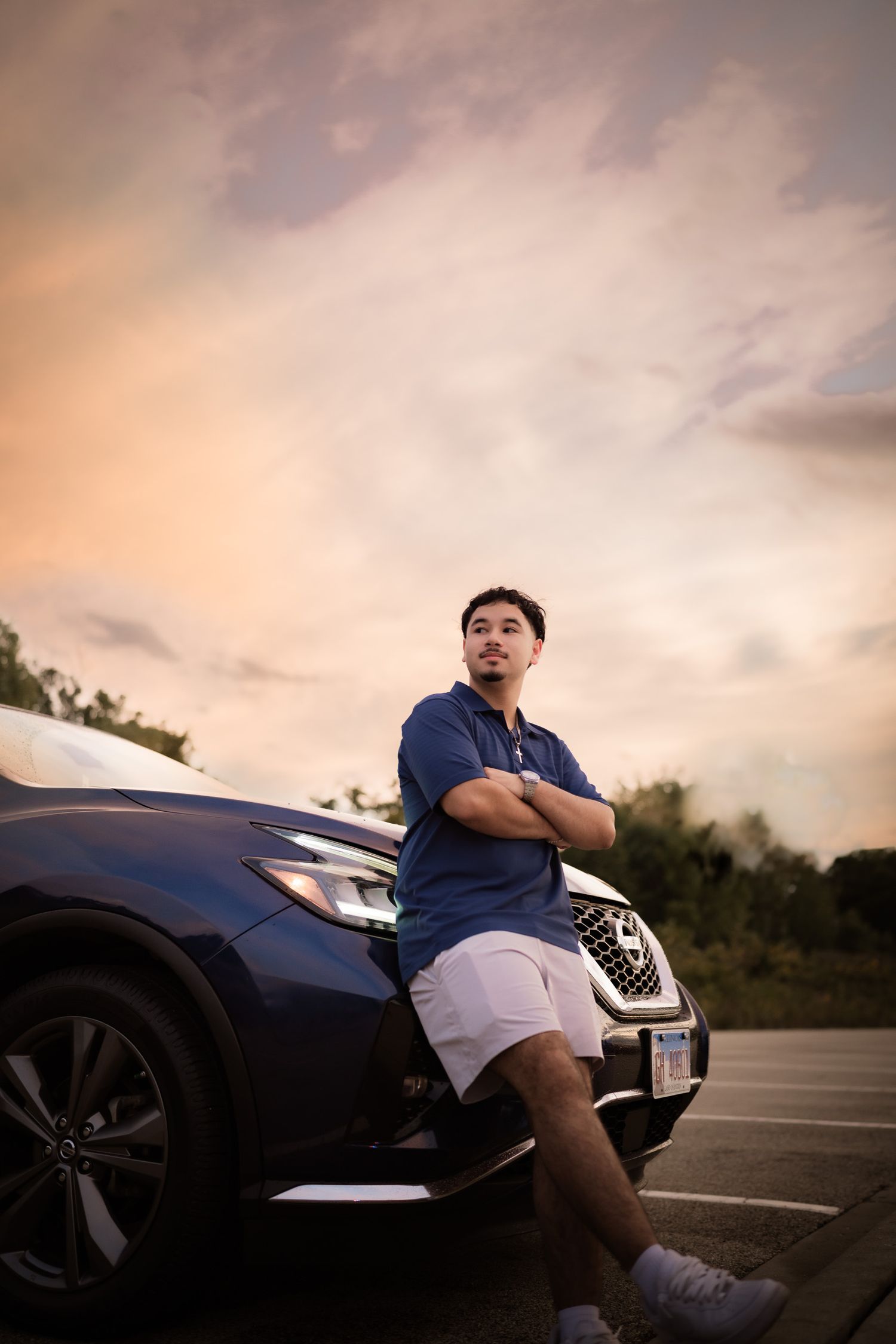 Man in blue shirt and white shorts leaning against a dark Nissan SUV in a parking lot at sunset.